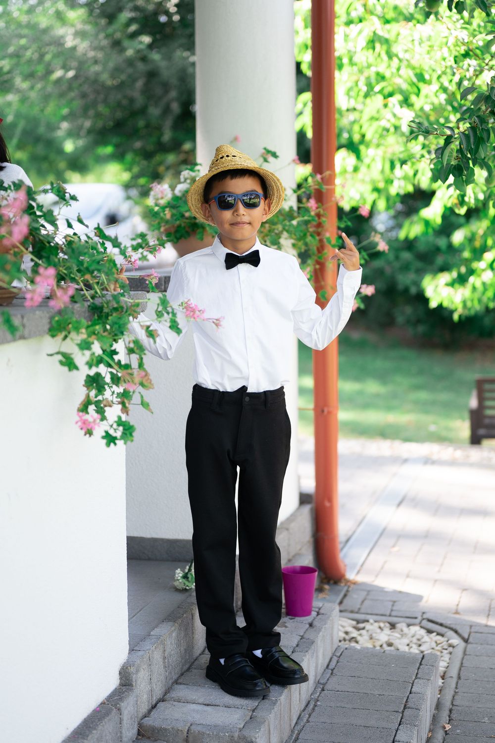 Stylish young boy in white shirt, bow tie, straw hat and sunglasses posing at a wedding.