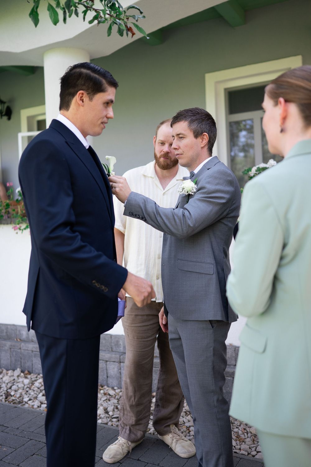 Groomsmen helping pin a boutonniere on a suit jacket outside before a wedding ceremony.