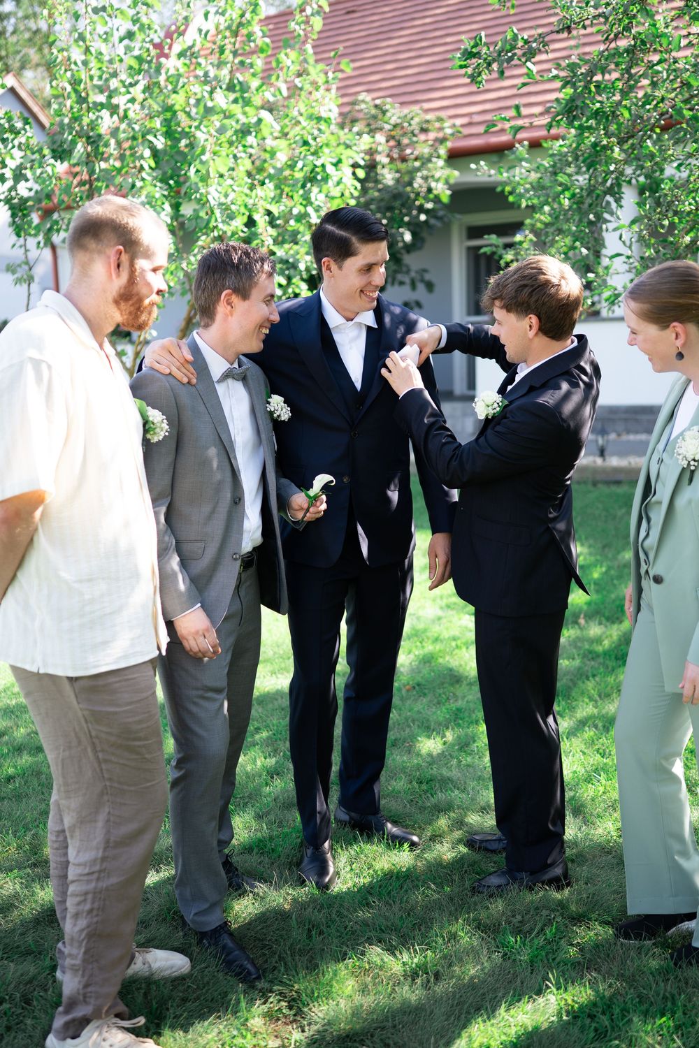 Groomsmen helping each other with boutonnieres outdoors at a wedding on a sunny day.