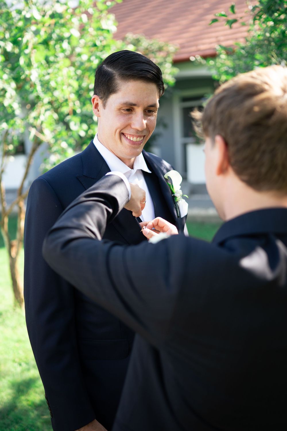 Smiling groom in black suit having his boutonniere pinned by a groomsman outdoors.