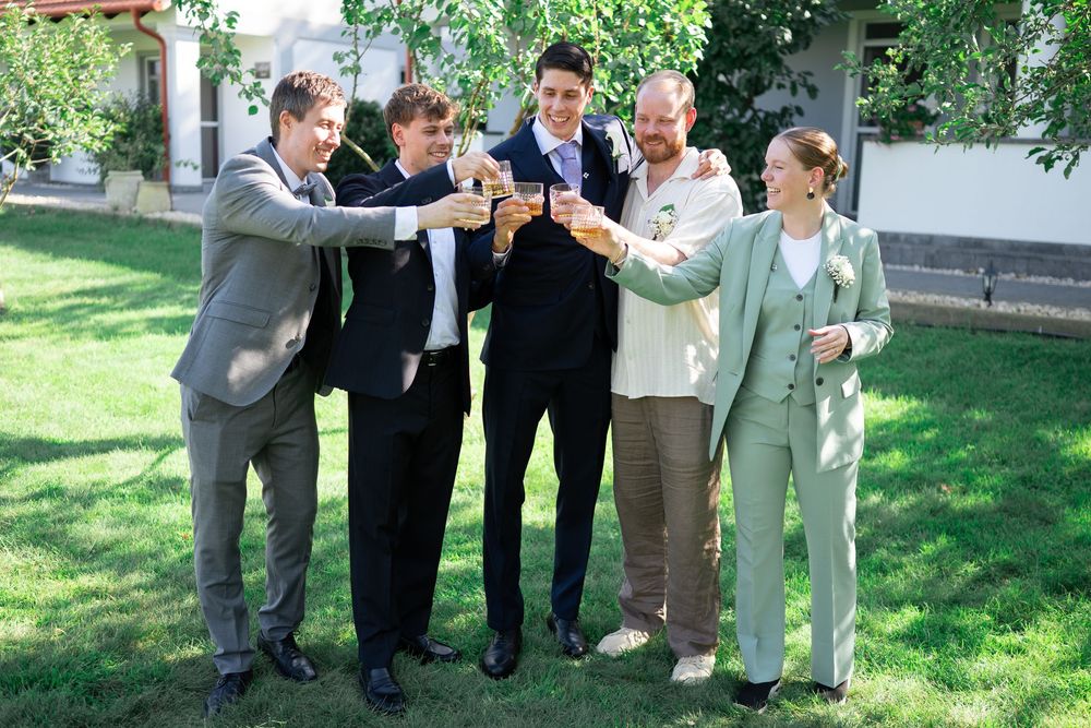 Group of five wedding party members toasting with drinks outdoors on a sunny day.