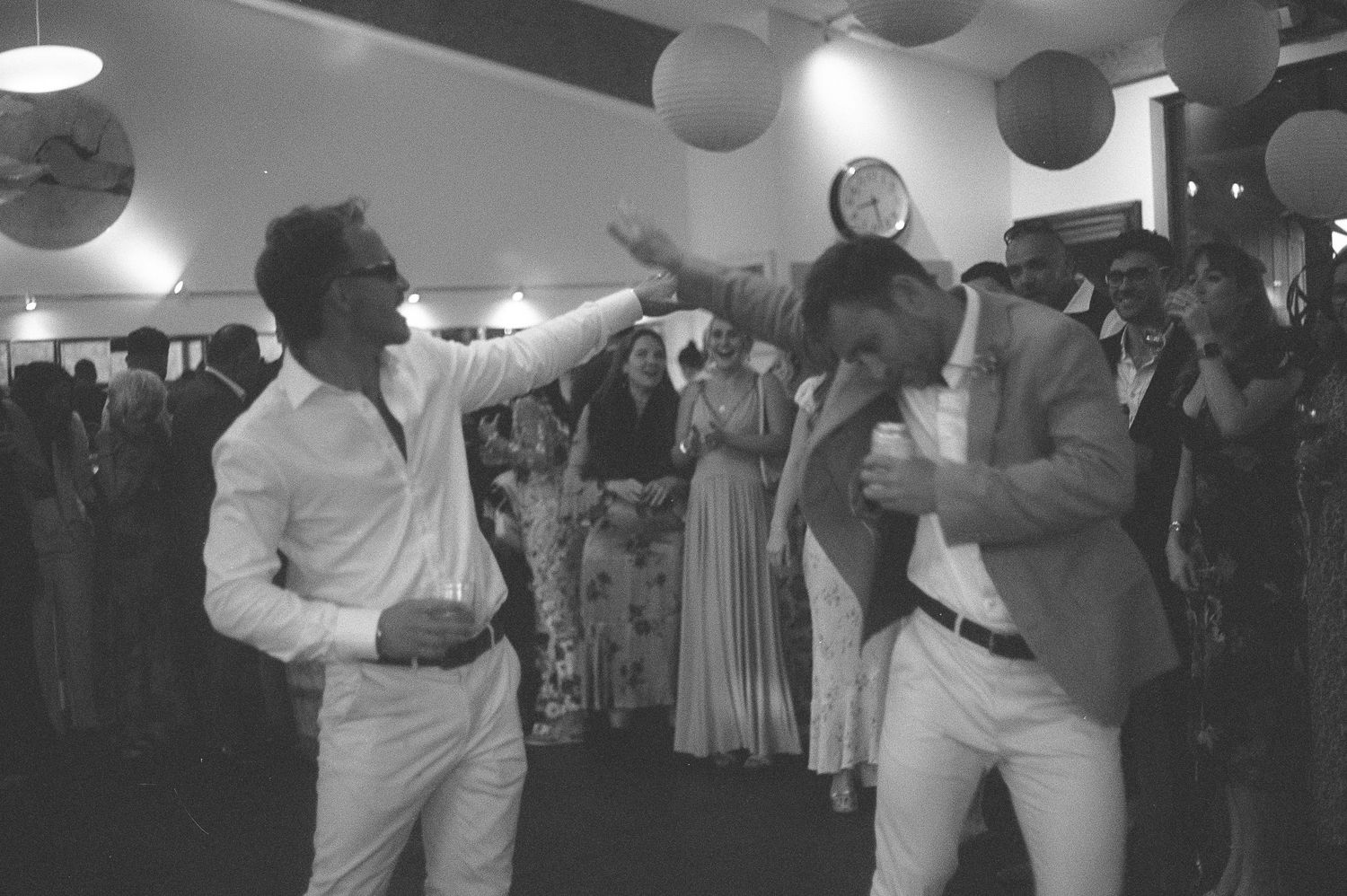 Black and white photo of two men dancing at a wedding reception with guests and decorative lanterns in the background.