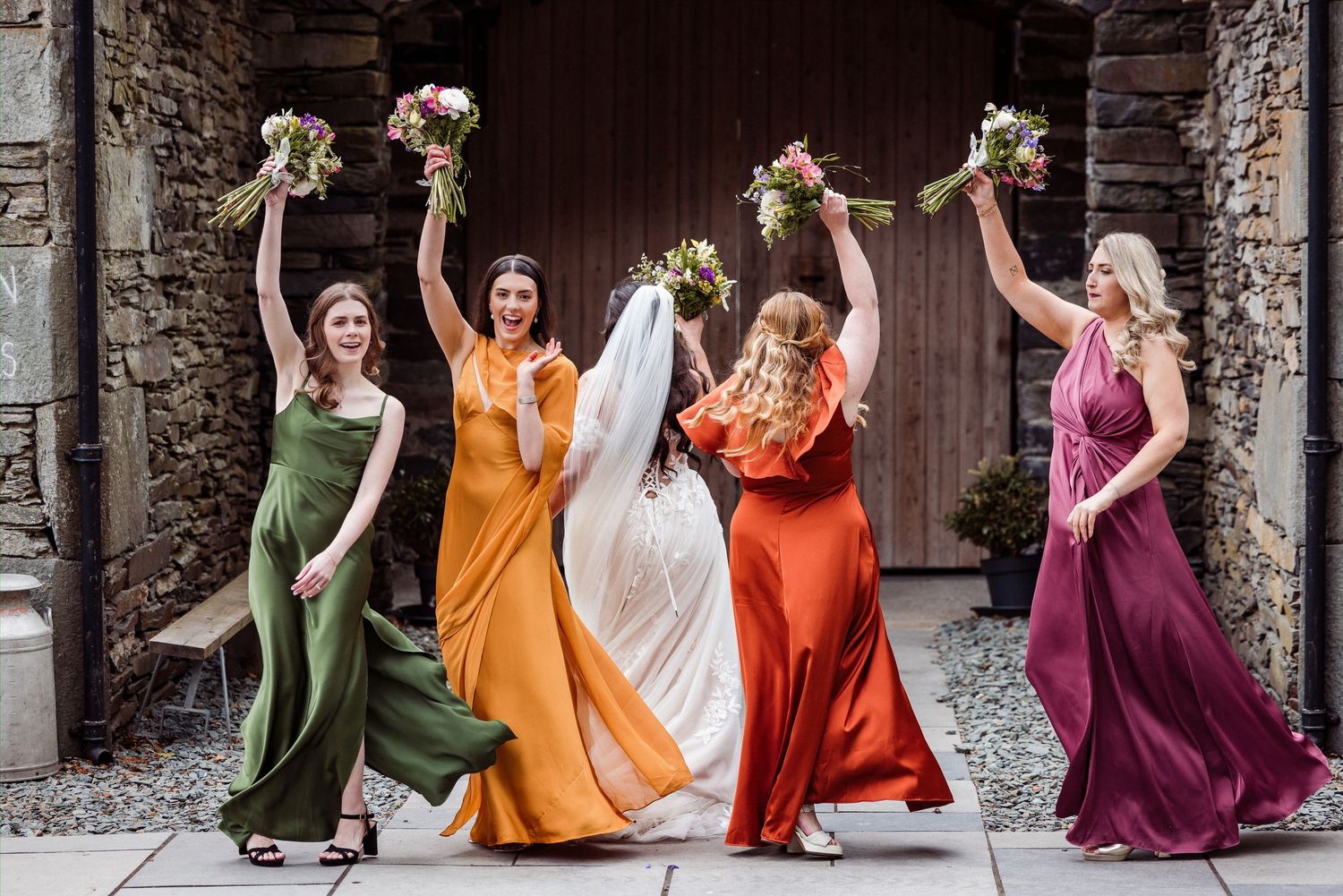 Bride and four bridesmaids in colorful gowns joyfully raising bouquets outside a rustic stone building.
