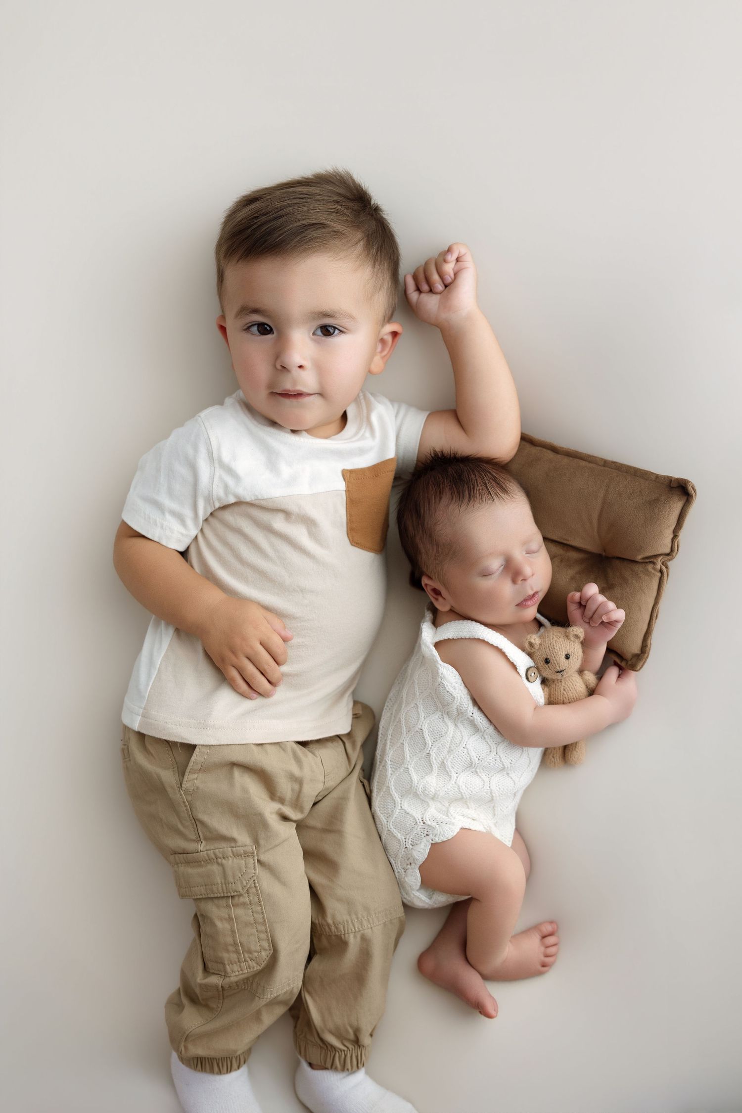 A toddler boy in khaki pants poses beside his sleeping newborn sibling holding a teddy bear on a white background.
