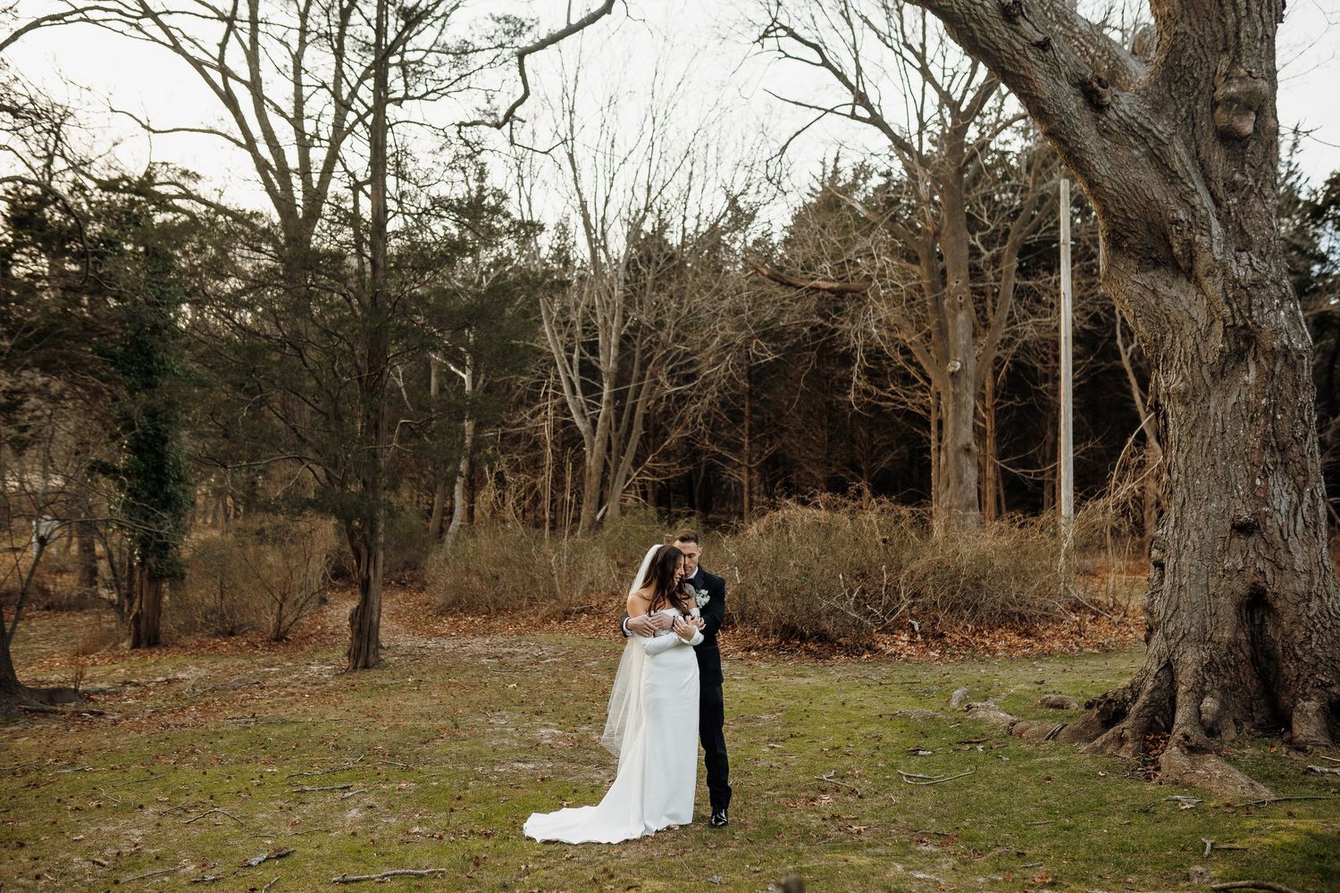 Bride and groom embracing near a large tree in a wooded outdoor setting during their wedding day.