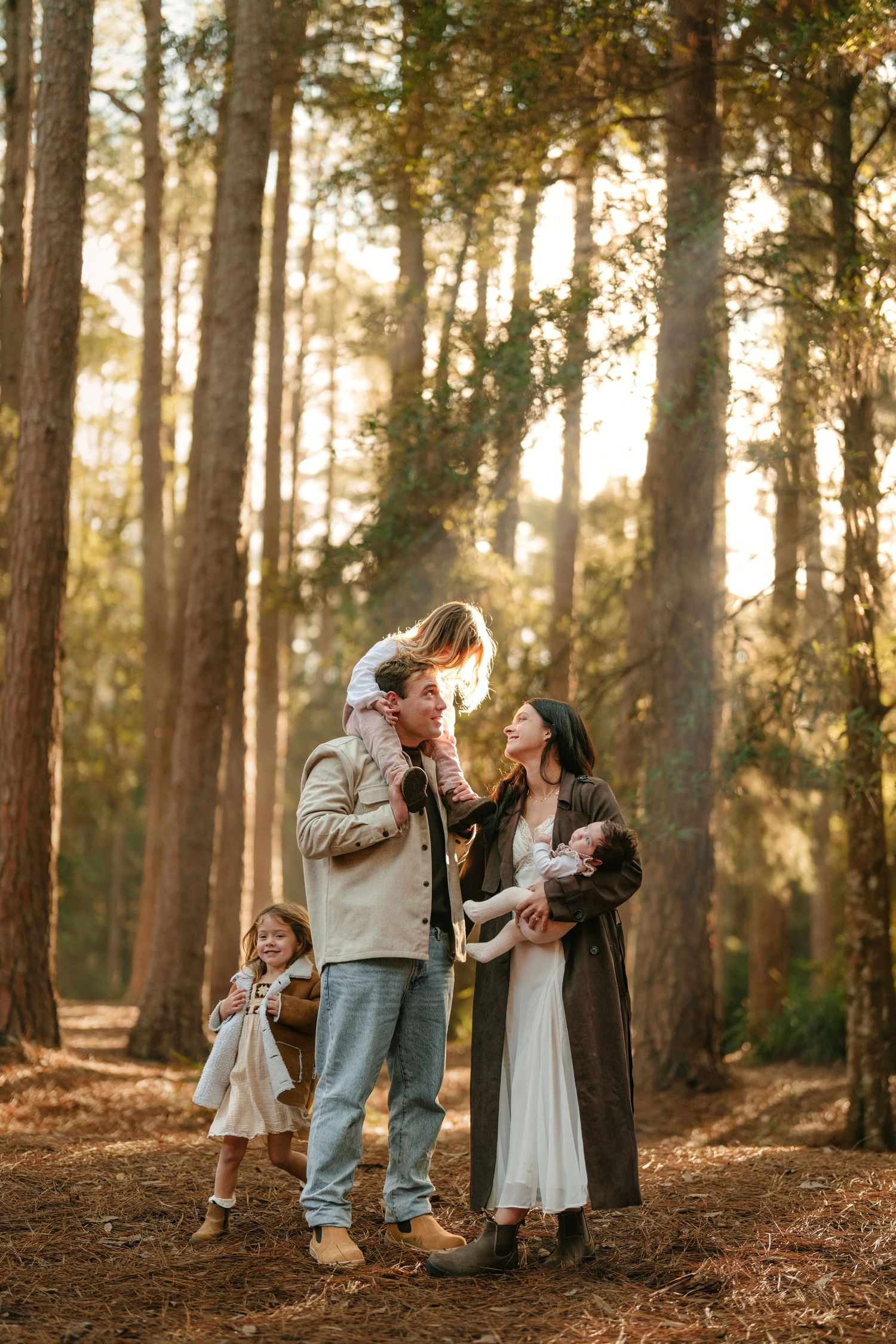 Family of four with newborn and two daughters standing in a sunlit pine forest at golden hour.