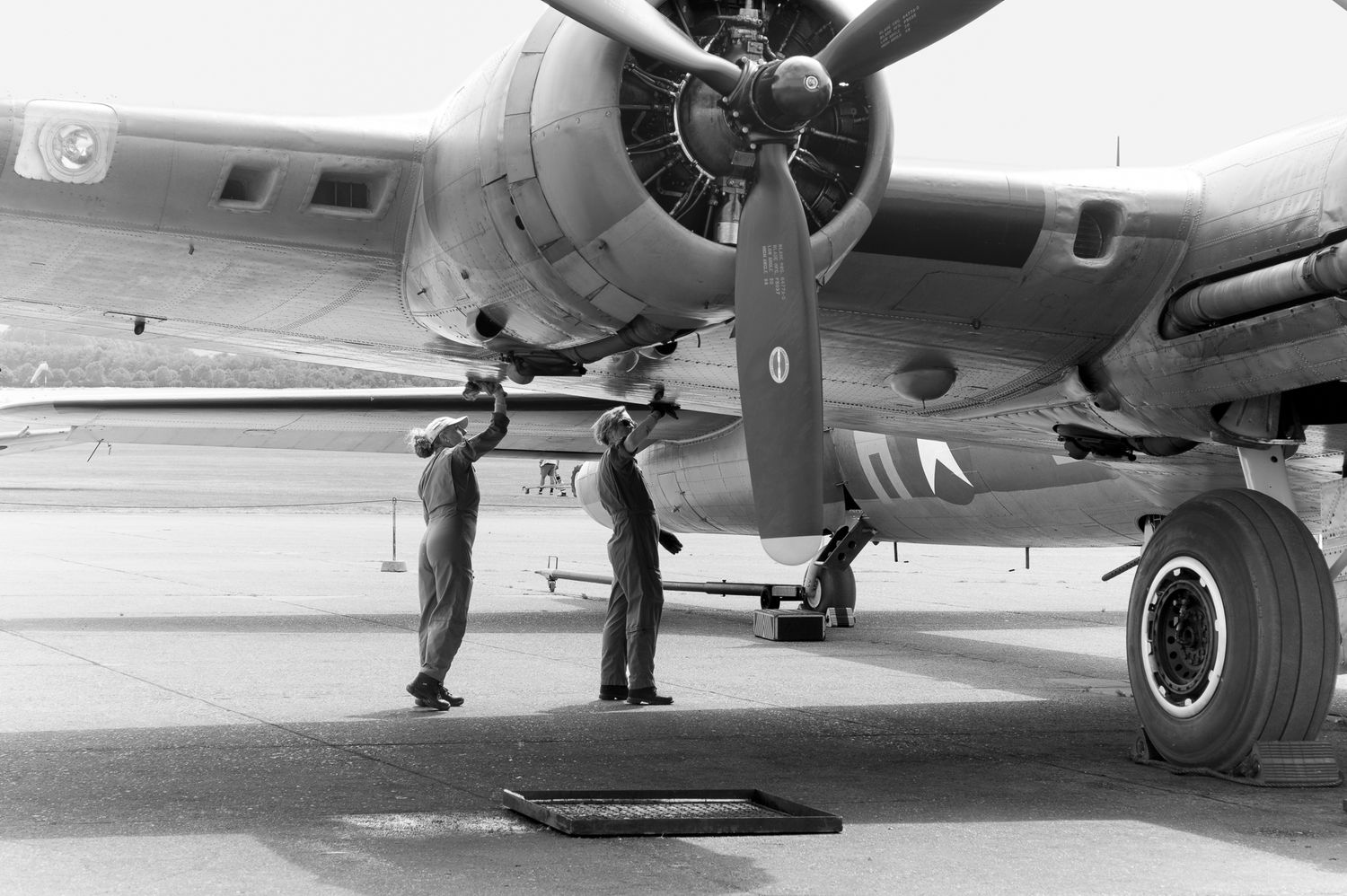 WWII-era bomber aircraft with checkered nose art being inspected by ground crew in vintage black and white photo.