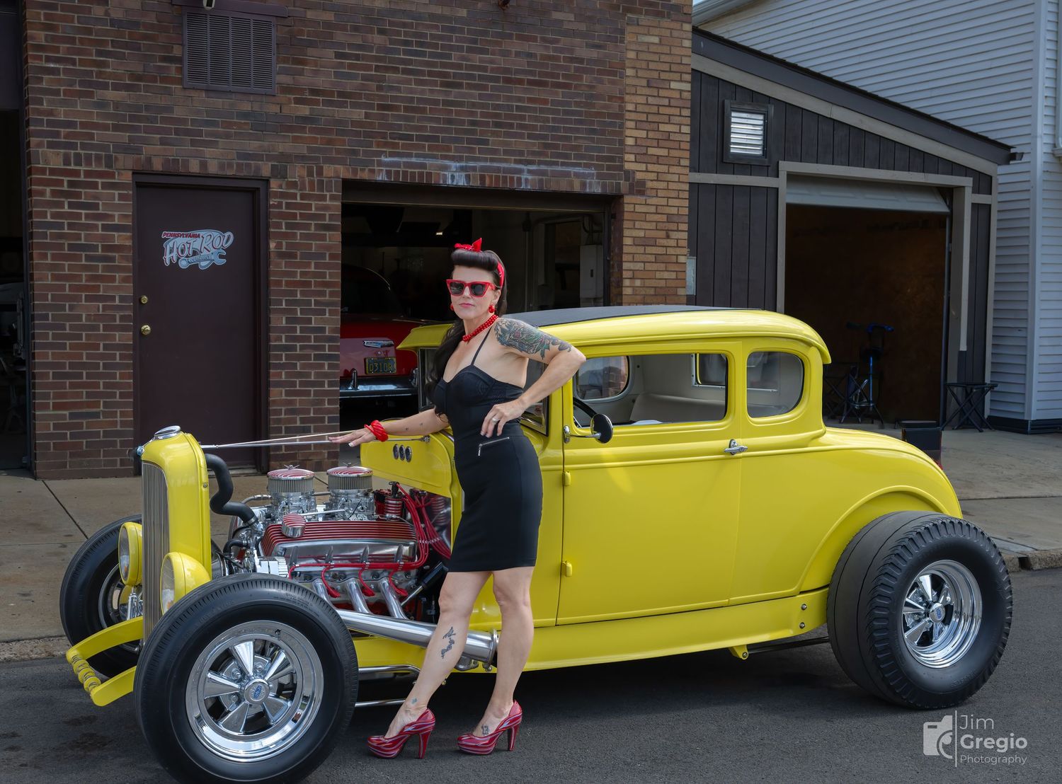 Woman in vintage pin-up style poses beside a yellow hot rod classic car outside a brick garage.