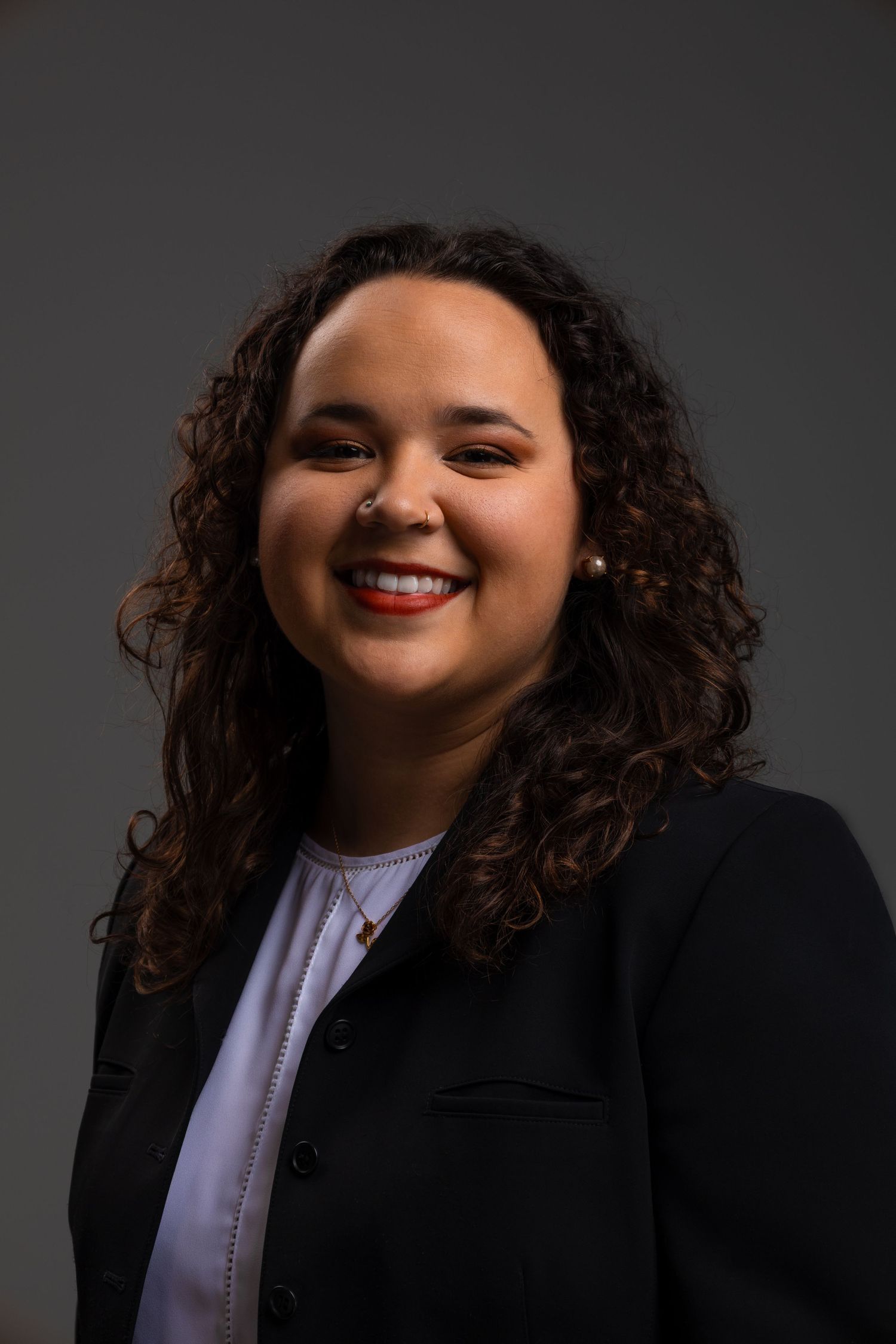 Professional headshot of a smiling young woman with curly hair wearing a black blazer and white top.