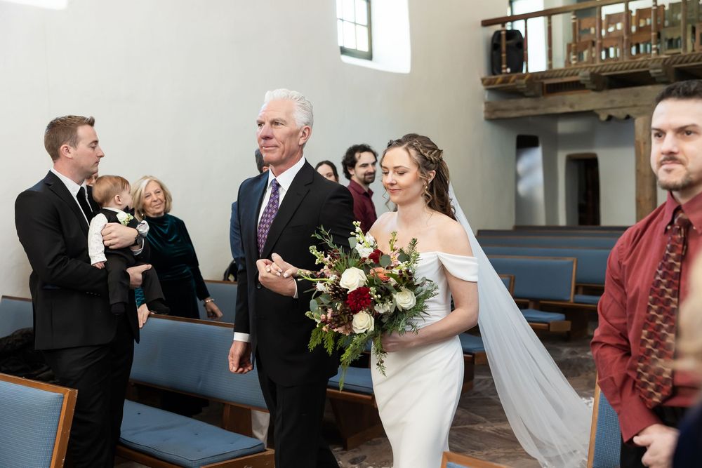 Bride walks down church aisle with father, holding floral bouquet, as guests watch during wedding ceremony.