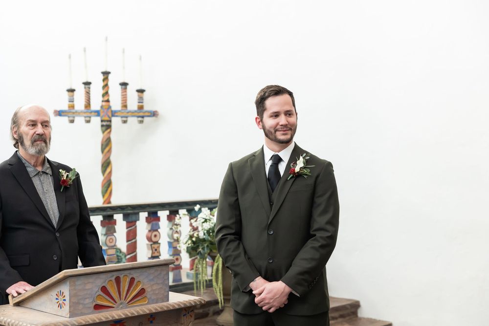 Two men in formal attire stand at a wedding altar, one in an olive suit with boutonniere, smiling warmly.