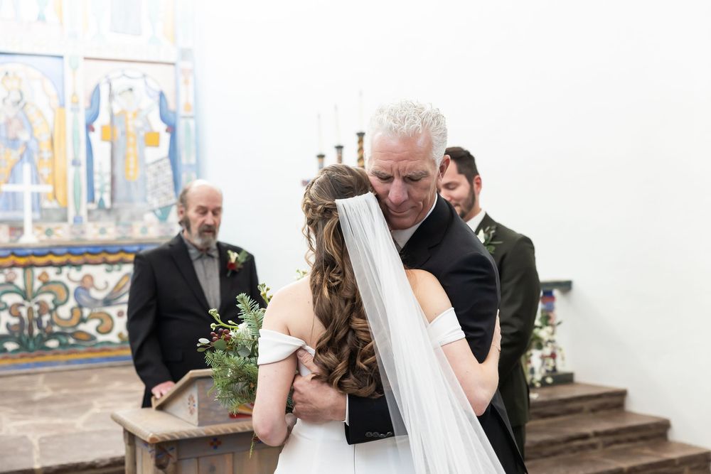 Father embracing bride in a church wedding ceremony, with guests and colorful stained glass windows in the background.