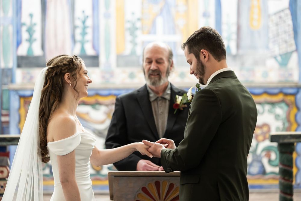 Bride and groom exchanging rings at altar during wedding ceremony with officiant and colorful stained glass backdrop.