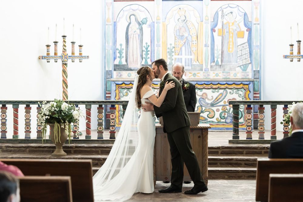 Bride and groom share first kiss at altar in a colorful church during wedding ceremony.