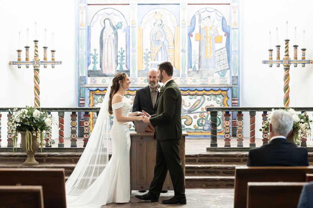 Bride and groom exchange vows at church altar with colorful religious mural backdrop during elegant wedding ceremony.