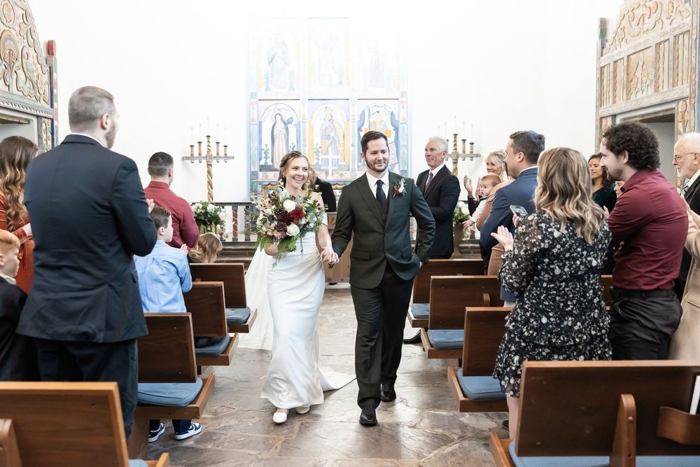 Bride and groom walk down church aisle after wedding ceremony, guests applauding in wooden pew-lined chapel.