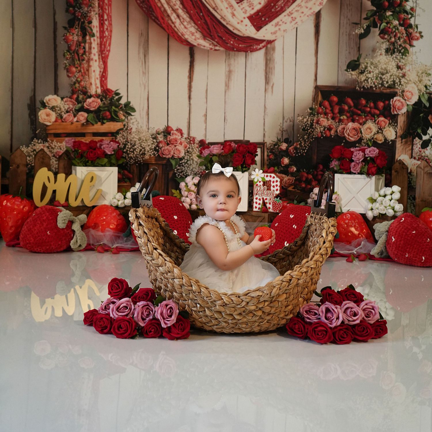 Baby girl in white dress sits in wicker basket surrounded by roses and strawberries at a floral first birthday cake smash.