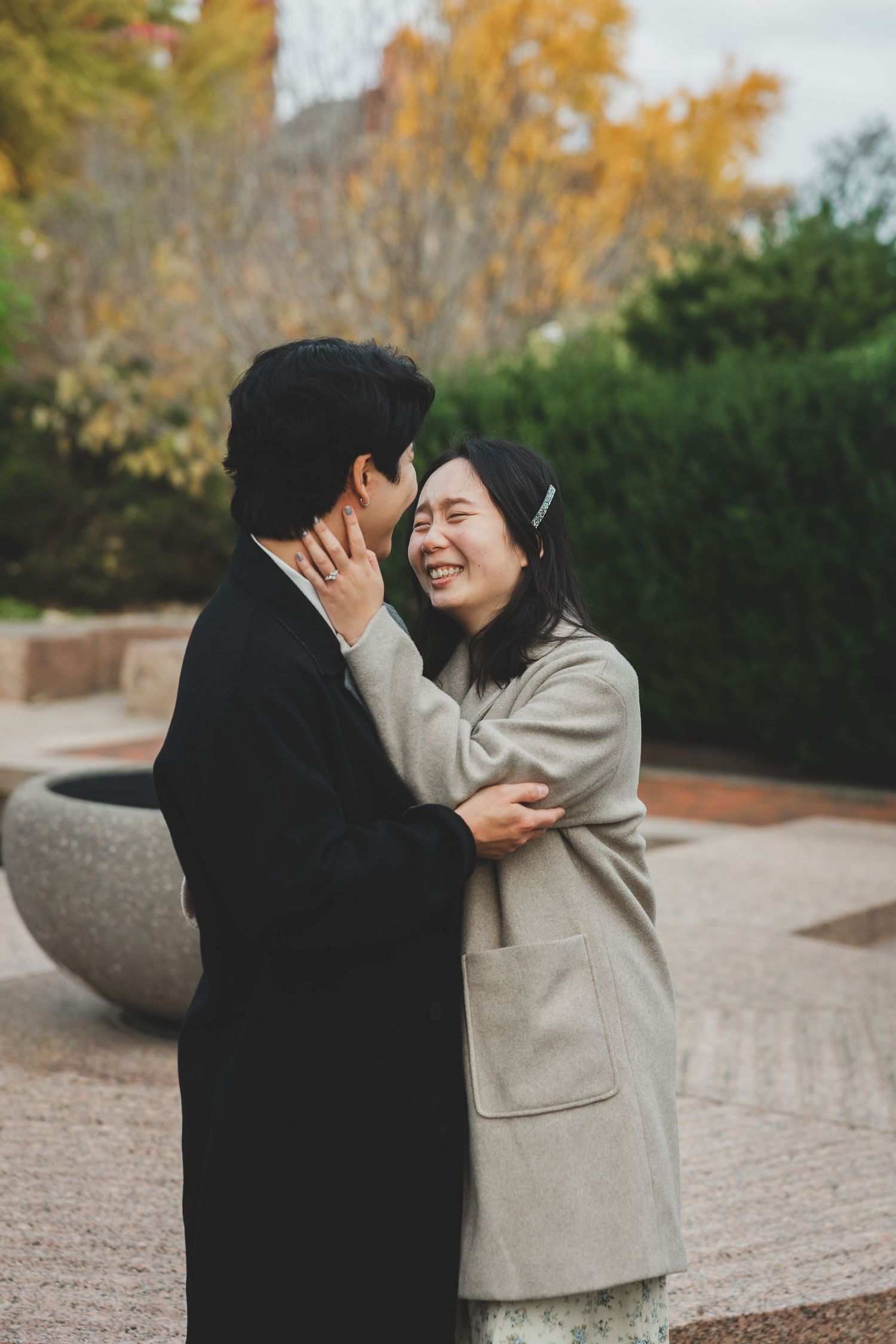 A joyful couple embraces outdoors in autumn, woman laughing as she touches mans face, both wearing elegant coats.