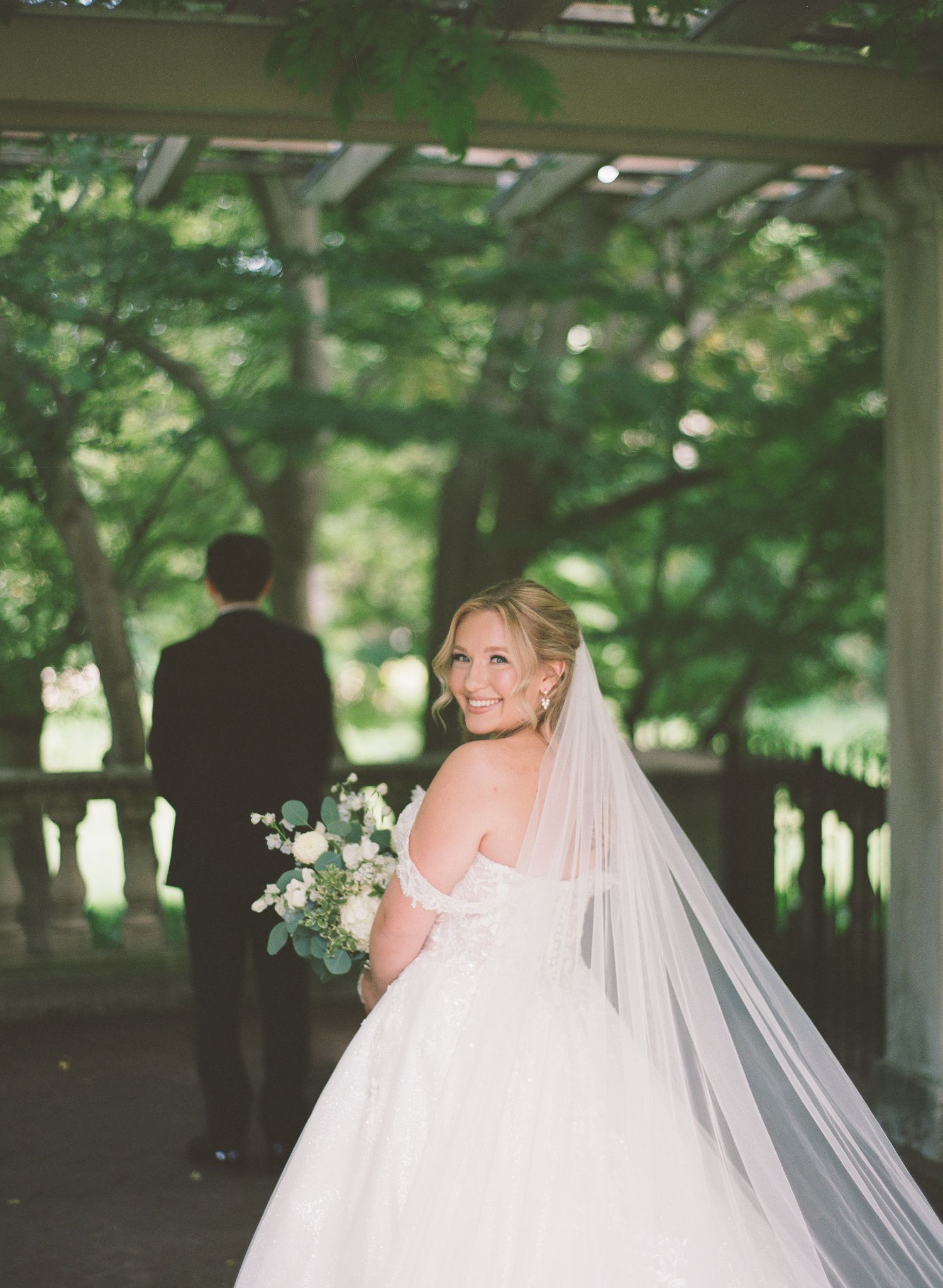Bride in off-shoulder gown with cathedral veil smiles back at groom under garden pergola.