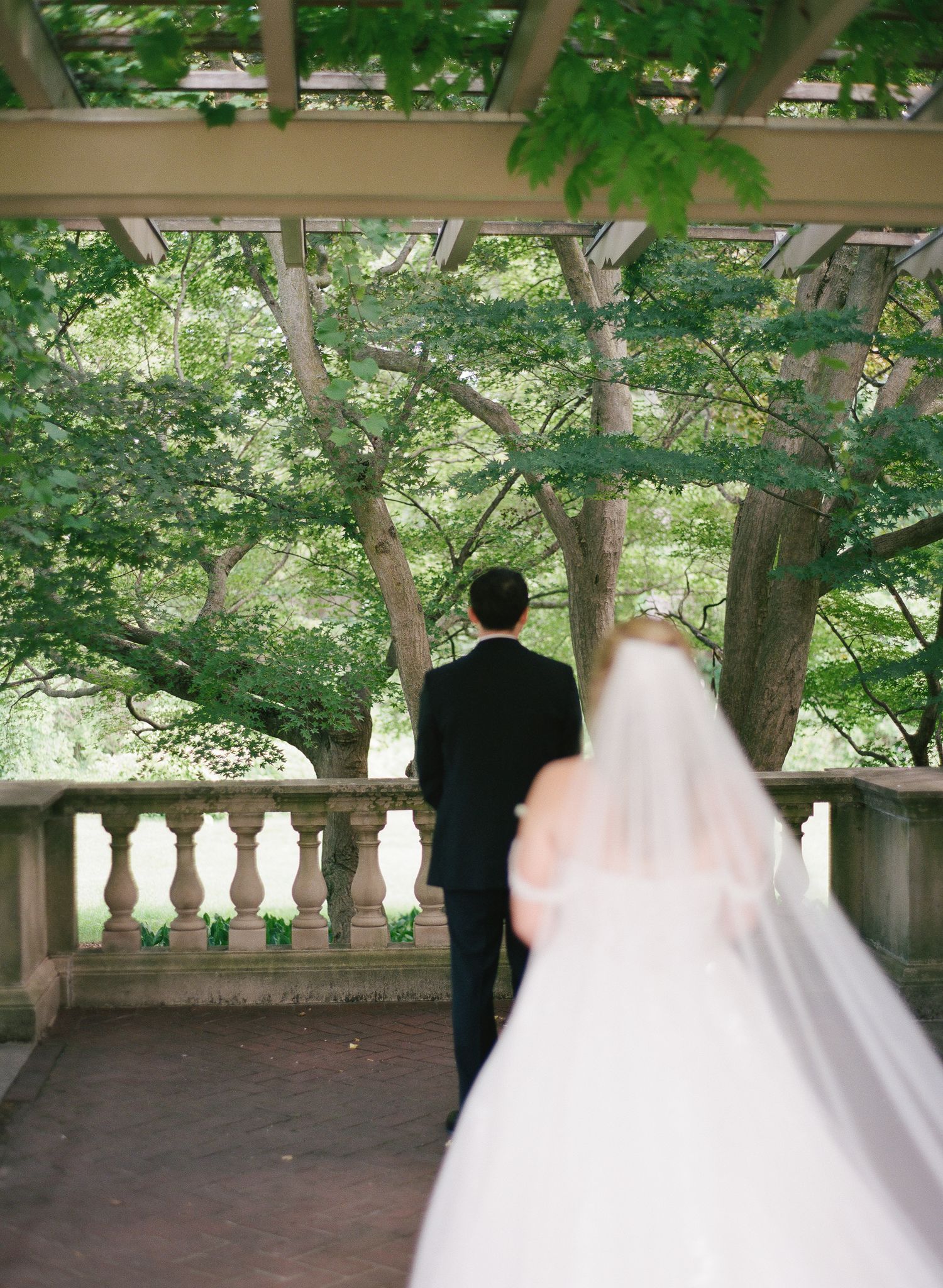 Bride and groom stand on an elegant stone balcony surrounded by lush green trees during their outdoor wedding ceremony.