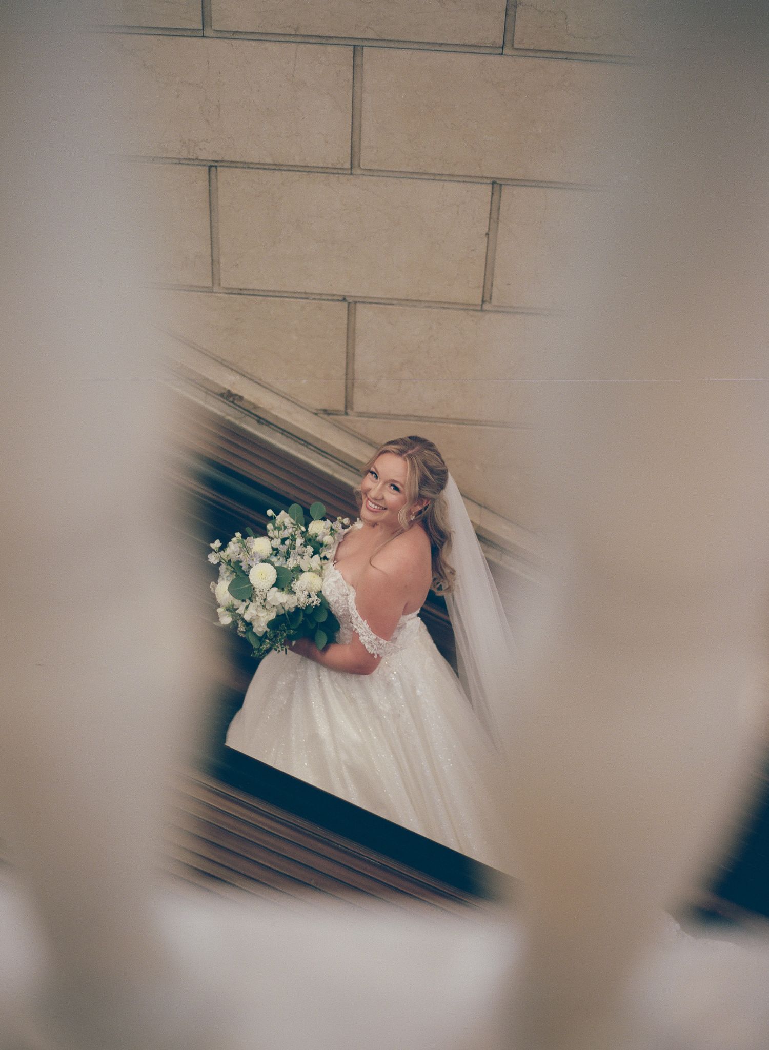 Bride in off-shoulder ball gown holding white bouquet with veil, smiling beside stone wall steps.
