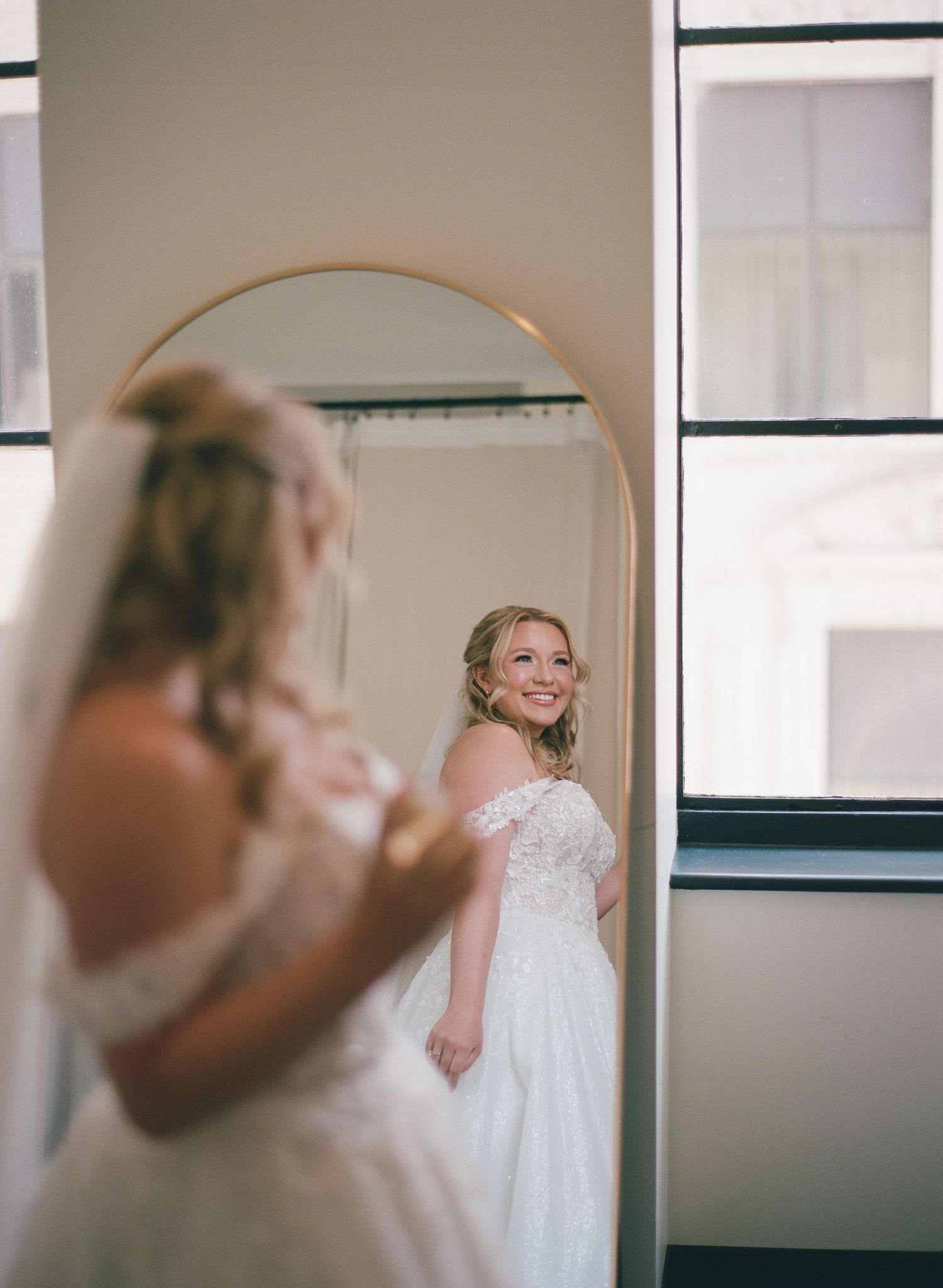 Smiling bride in off-shoulder lace wedding dress and veil admires her reflection in an arched gold mirror.