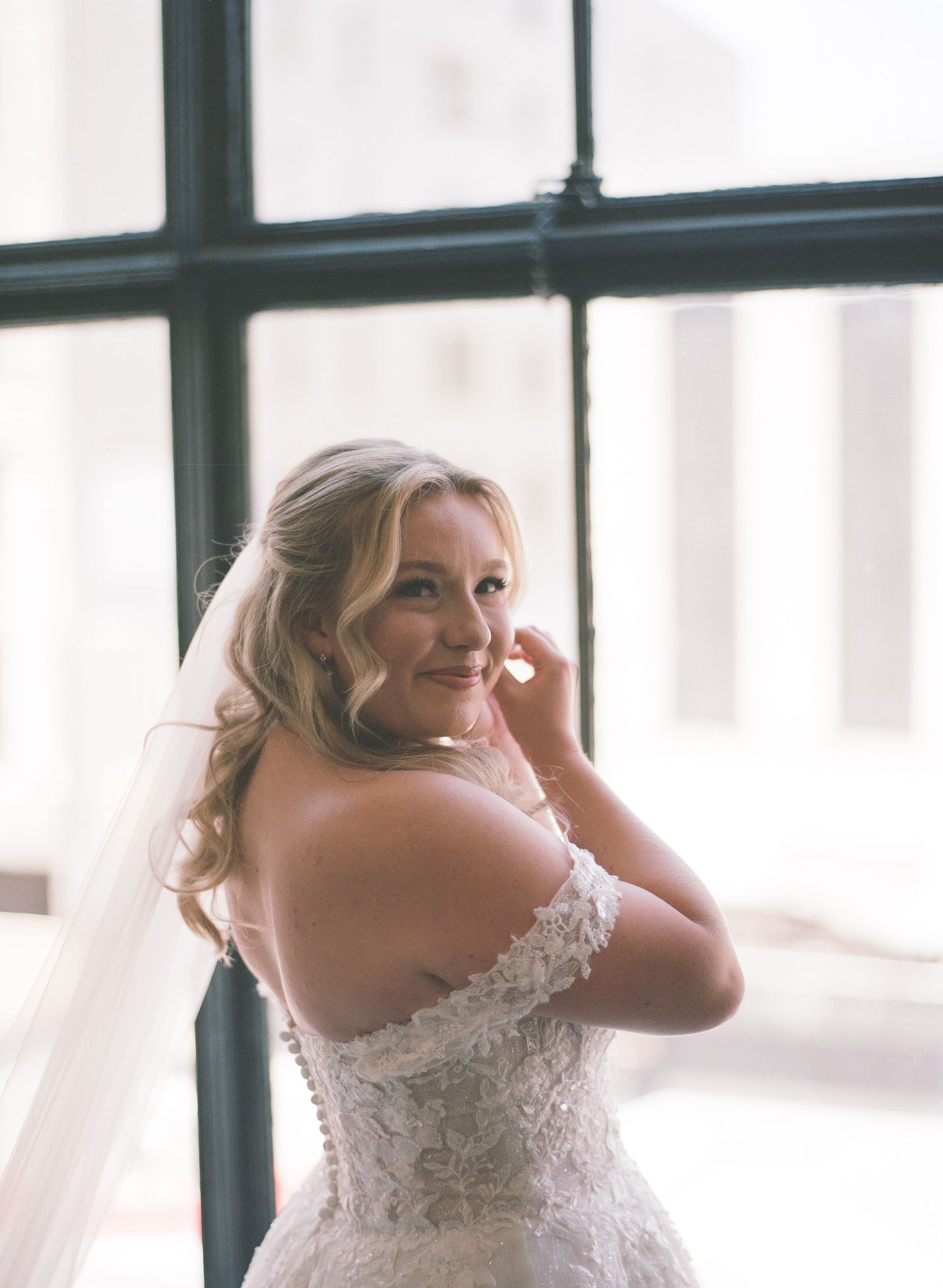 Bride in lace off-shoulder wedding gown and veil smiles over shoulder near large windows in soft natural light.