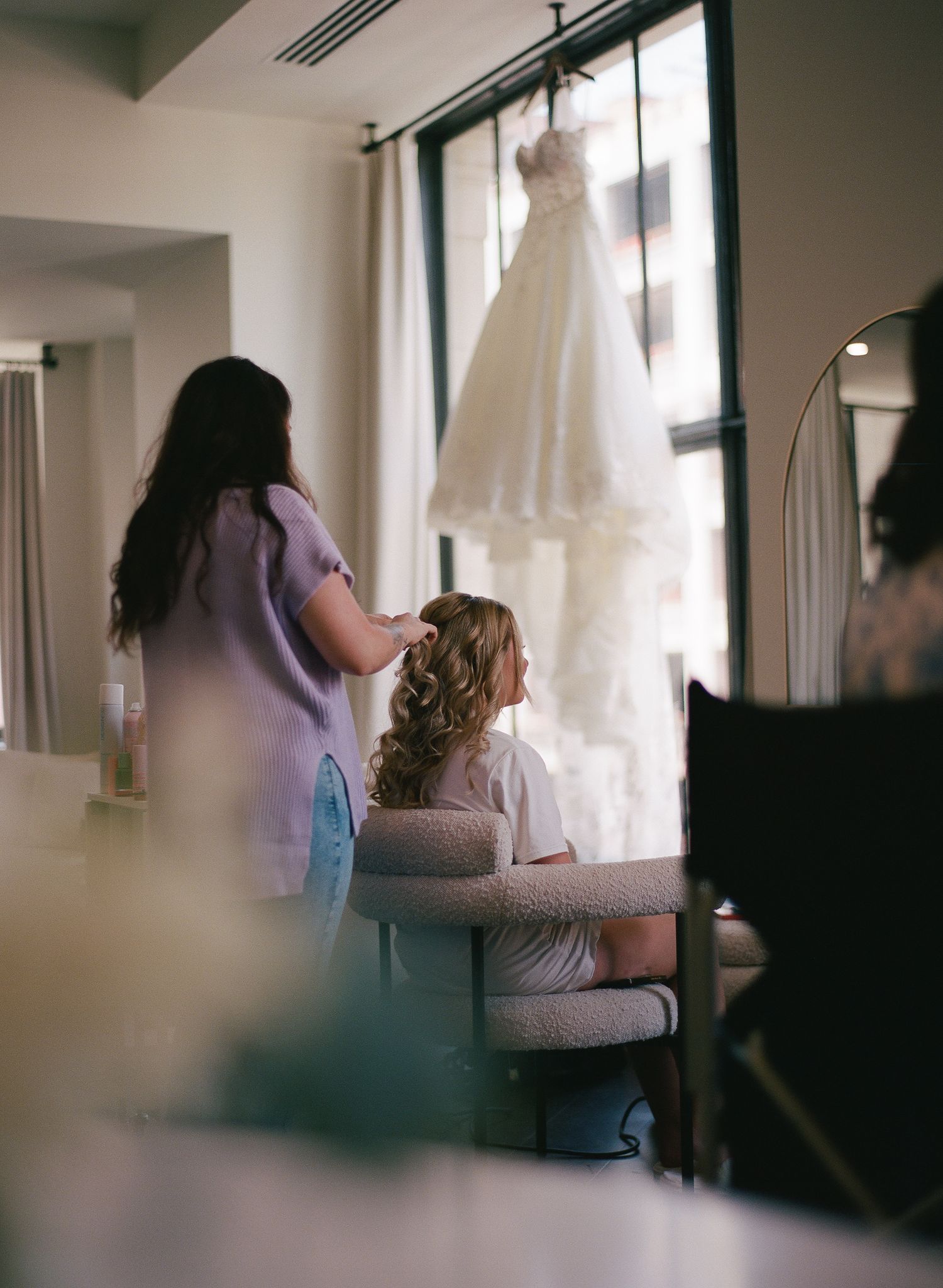 Bride getting hair done with wedding dress hanging in window during bridal preparations.