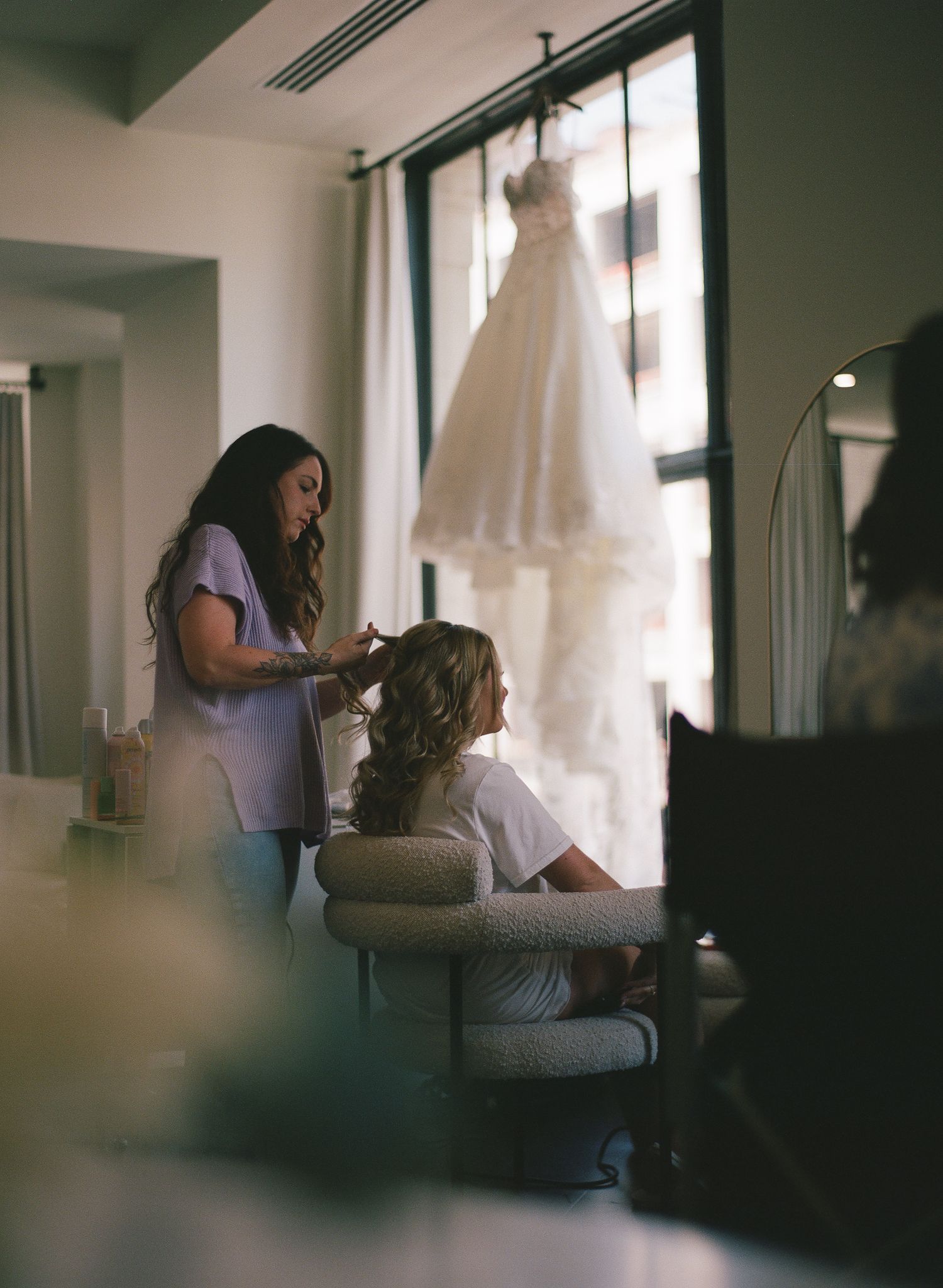 Bride getting hair styled in elegant room with wedding dress hanging in window background.