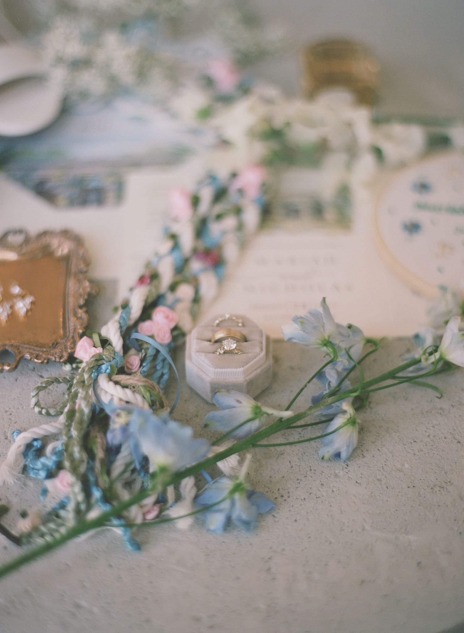 Elegant wedding ring in a velvet box surrounded by delicate blue and pink flowers on a styled flat lay.