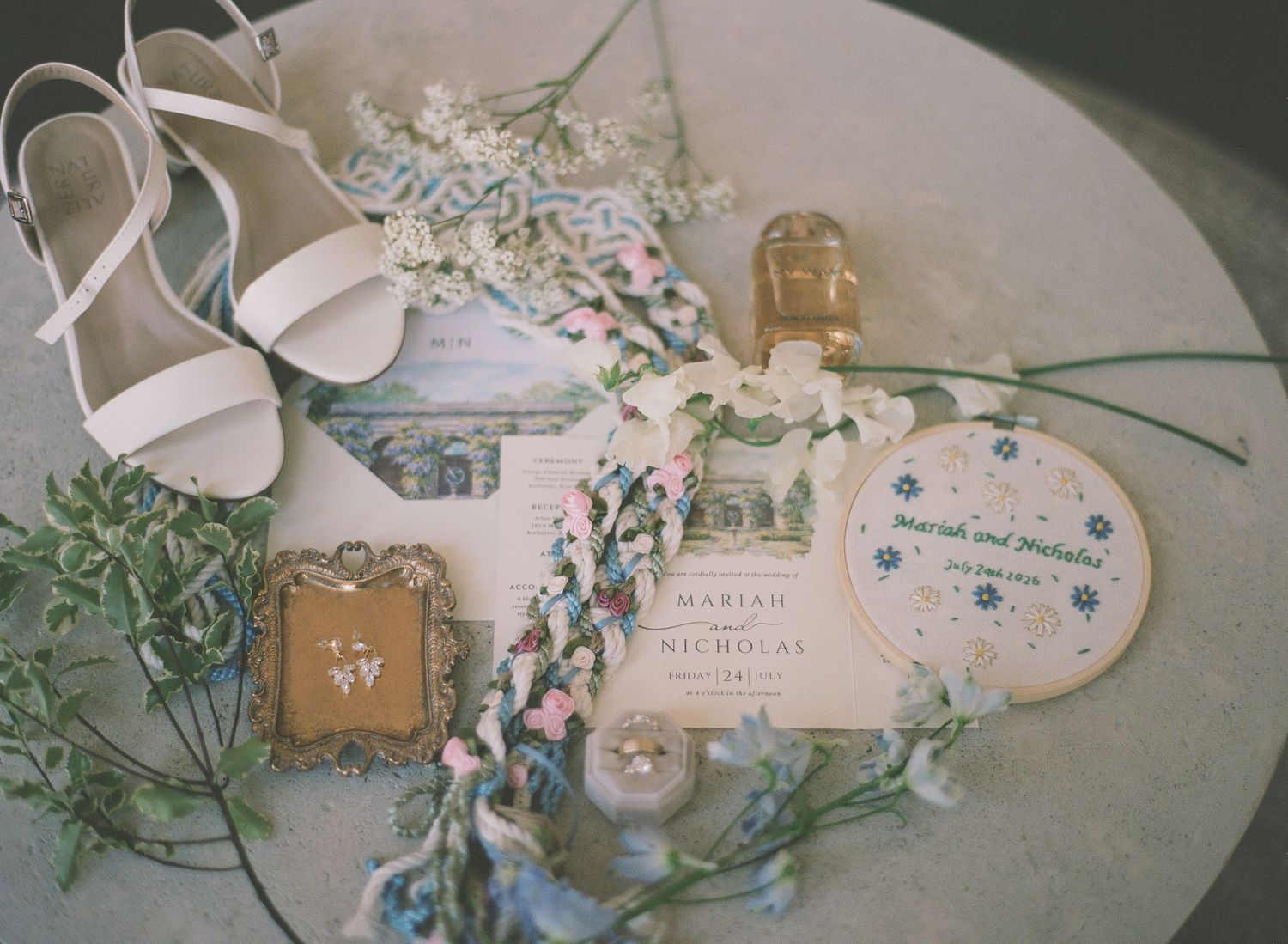 Wedding flat lay with white heels, floral garland, invitation suite, perfume bottle, and vintage gold frame on marble.
