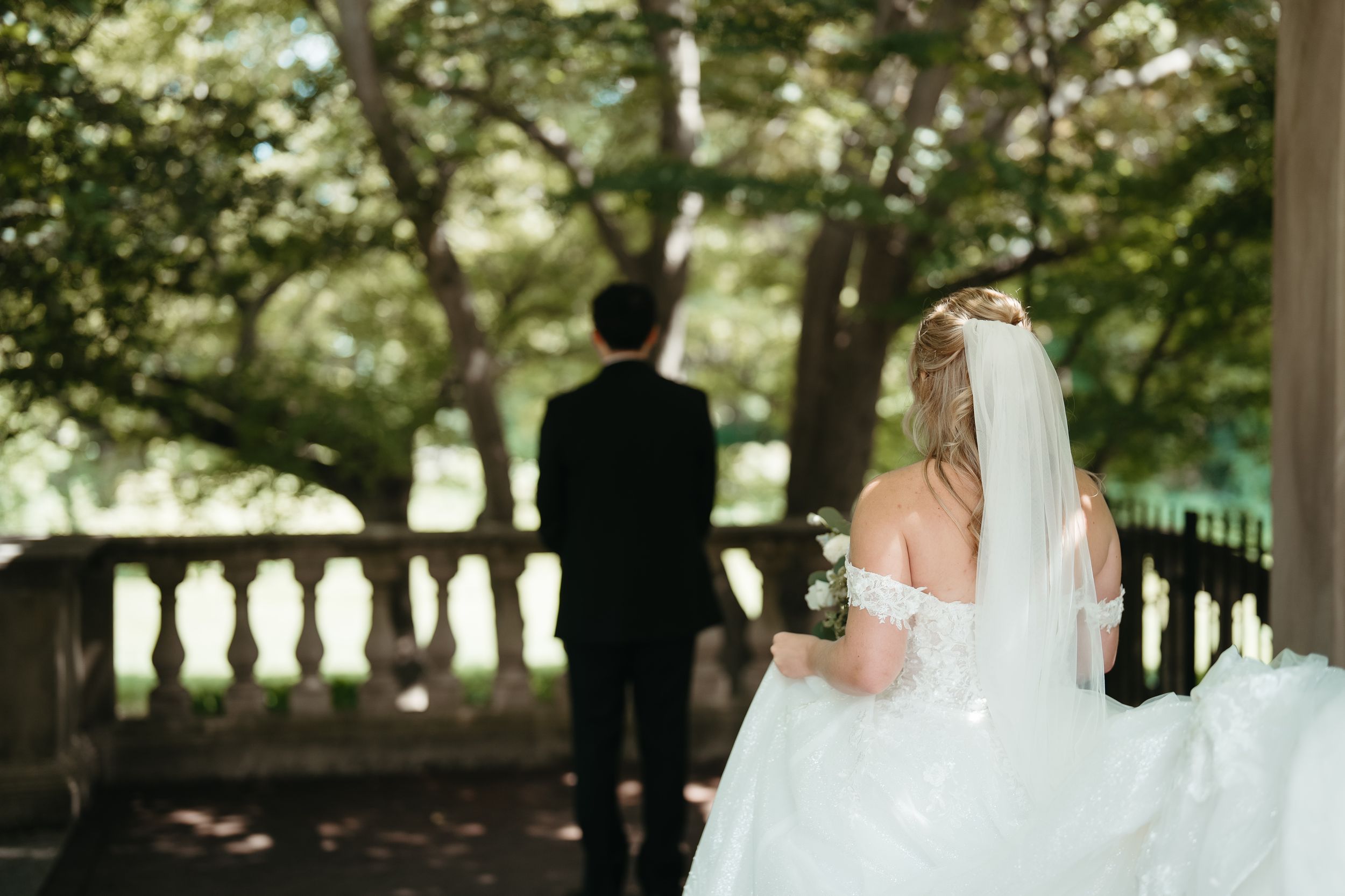Bride in white gown and veil walks toward groom in black suit at outdoor wedding venue surrounded by lush trees.