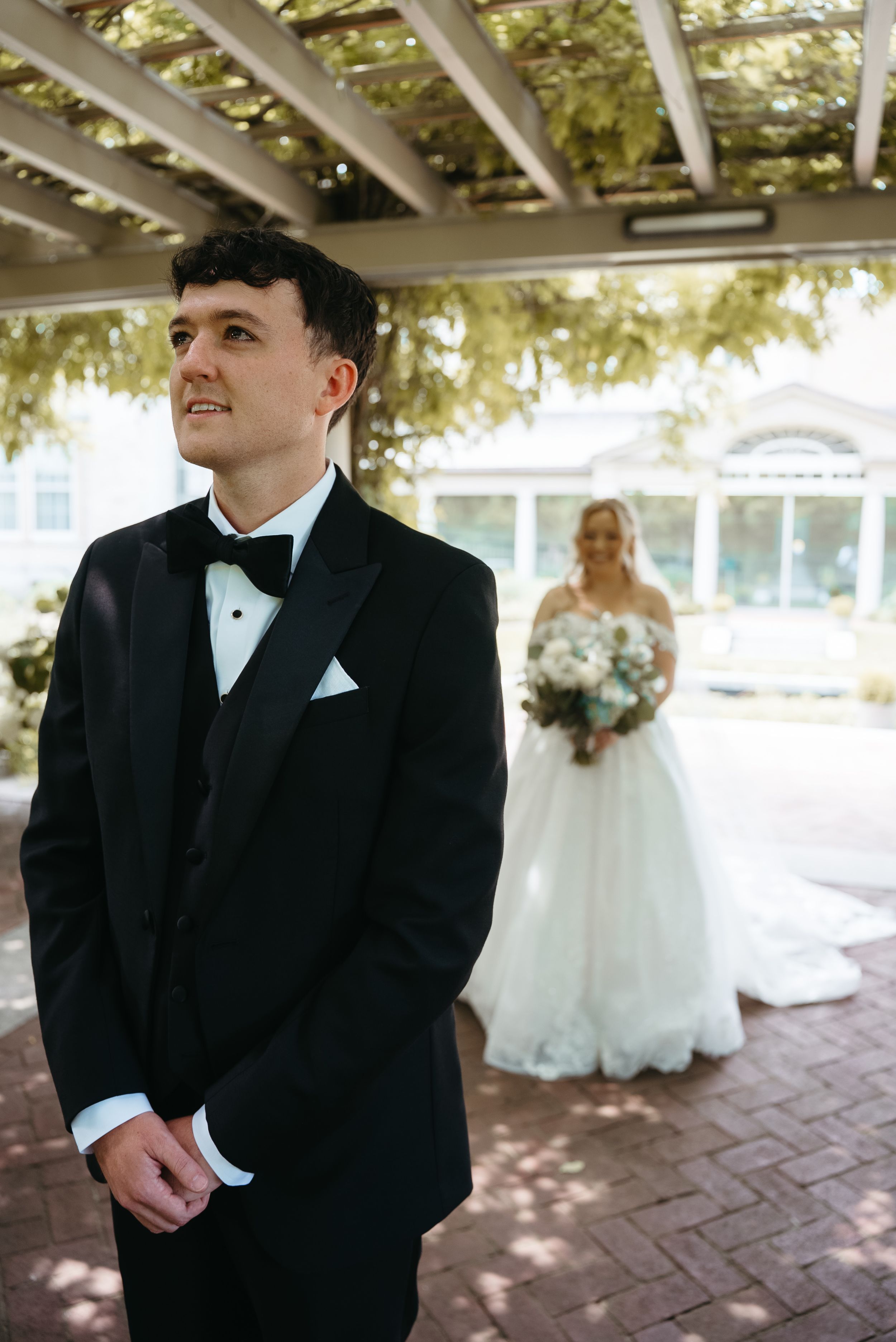 Groom in black tuxedo stands under pergola as bride in white gown holds bouquet in soft focus background.