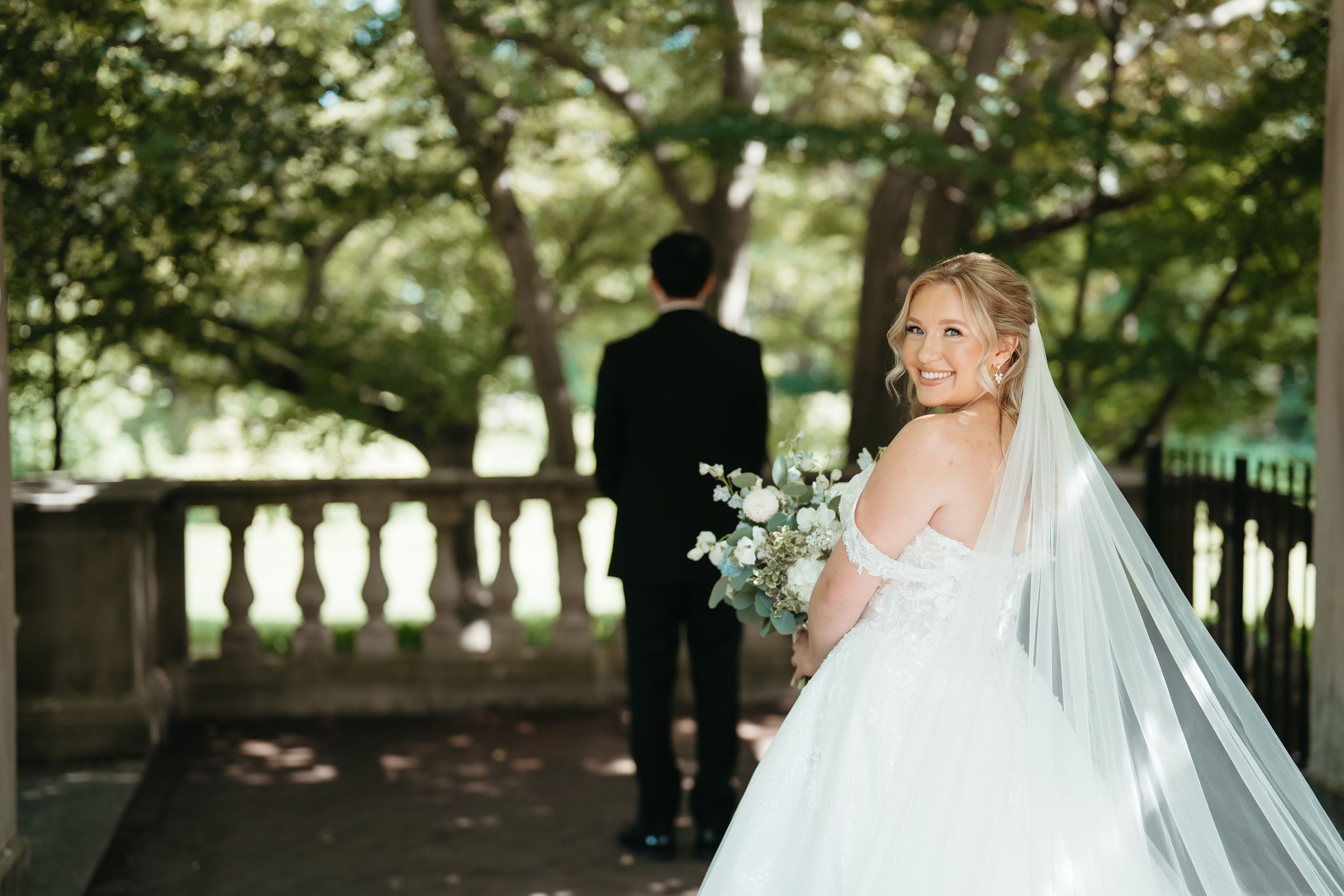 Bride in white gown and veil holds bouquet, smiling over shoulder, groom in black suit stands ahead on garden terrace.