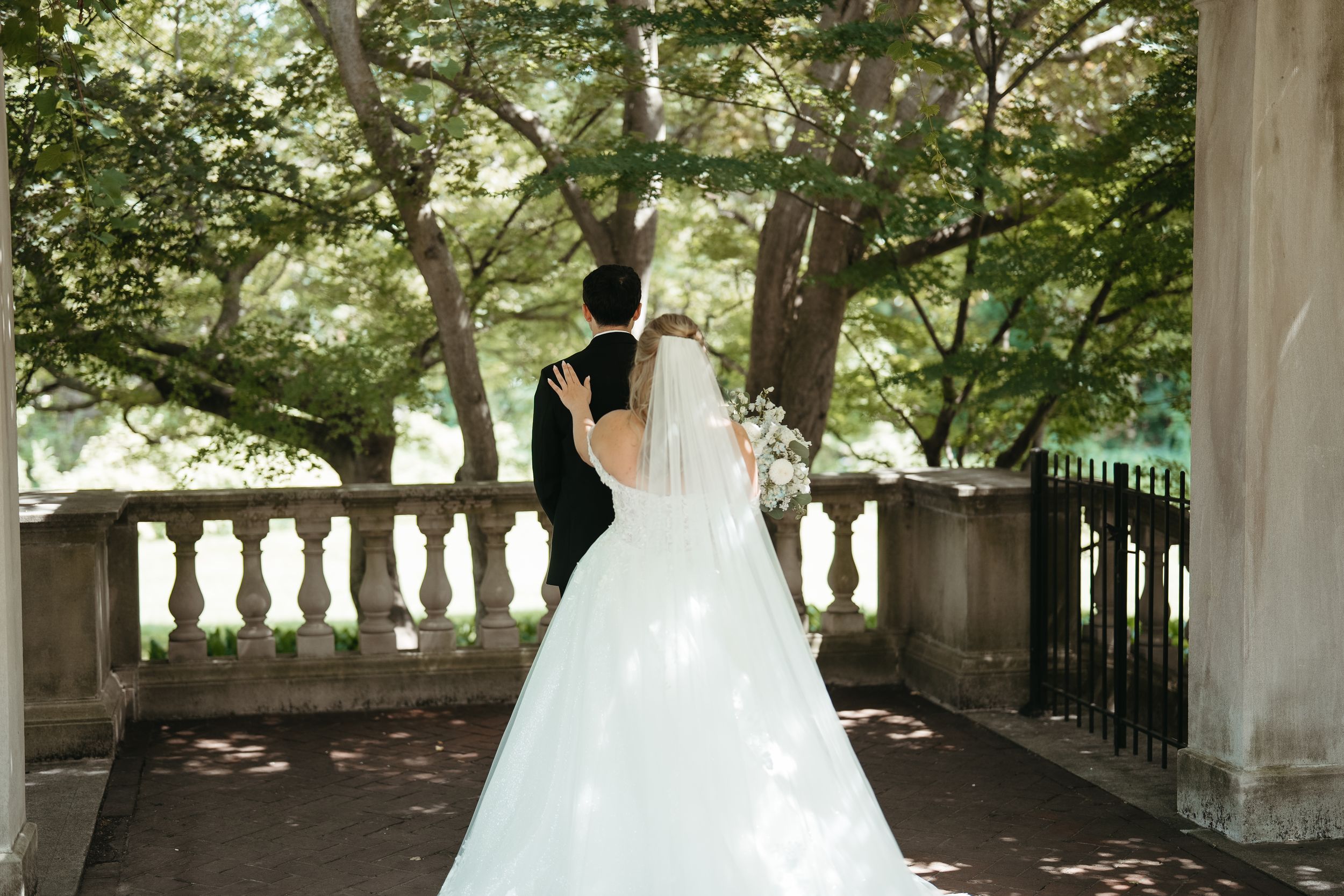 Bride and groom stand together on a stone balcony surrounded by lush green trees in an elegant outdoor wedding setting.