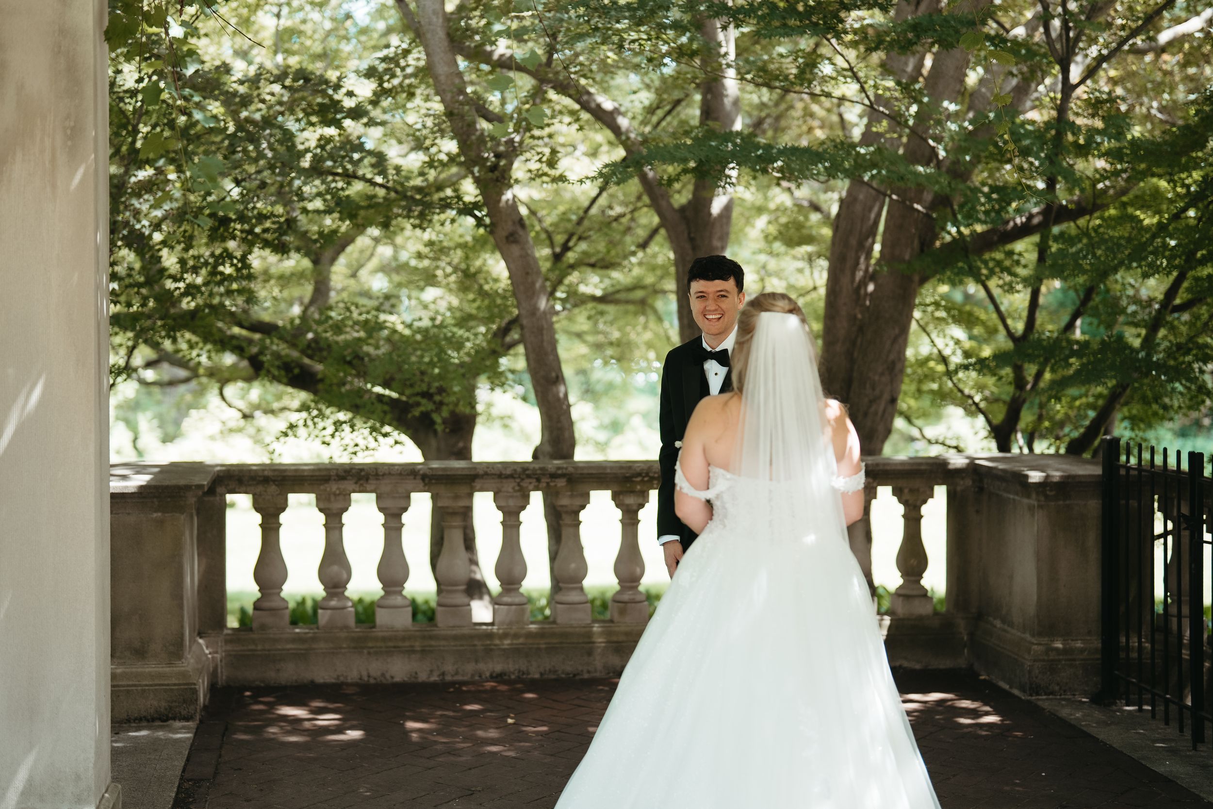 Bride in white gown and veil holds groom's hand in black tuxedo on stone balcony surrounded by lush trees.