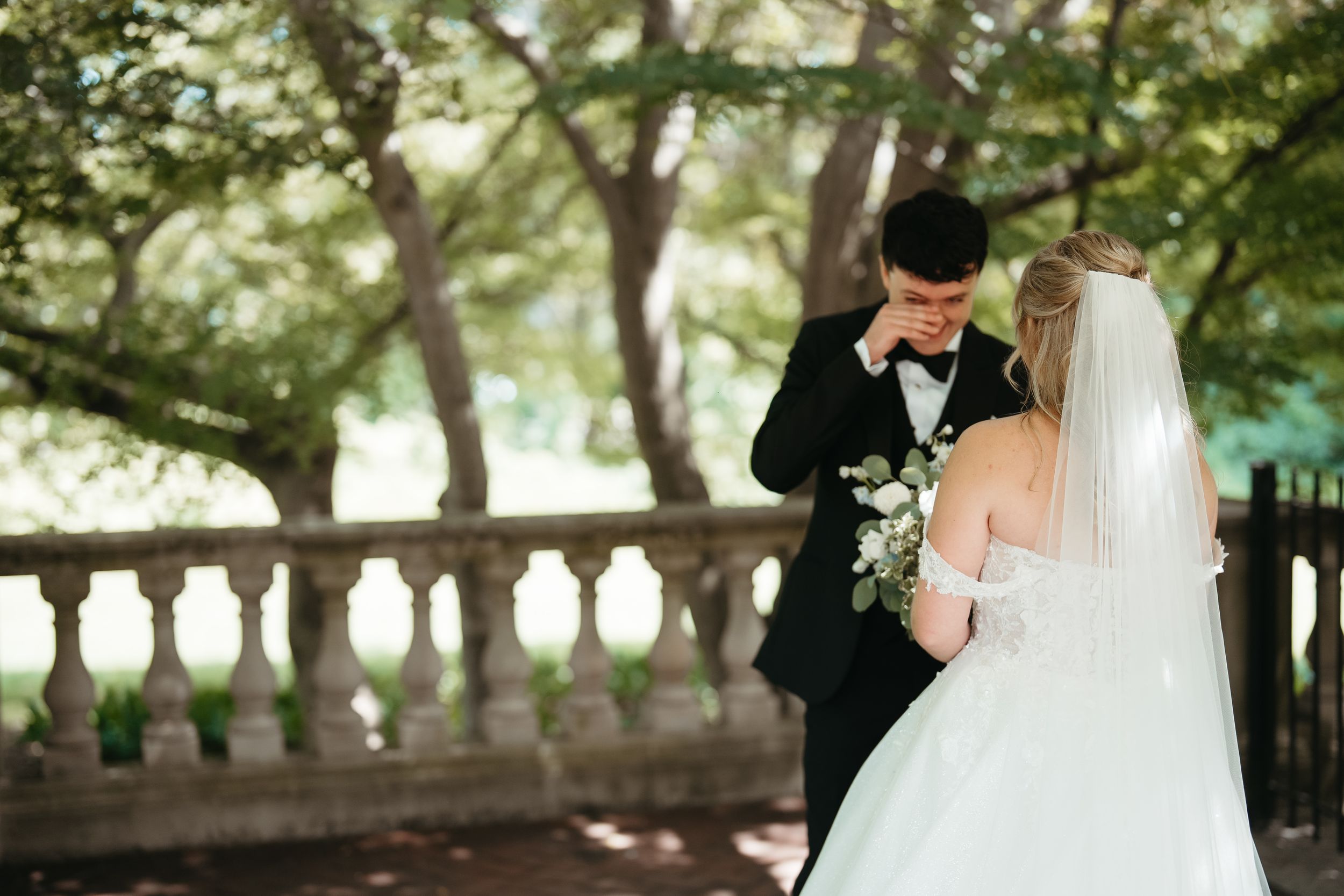 Groom wiping tears during emotional first look with bride in white gown and veil holding bouquet outdoors.