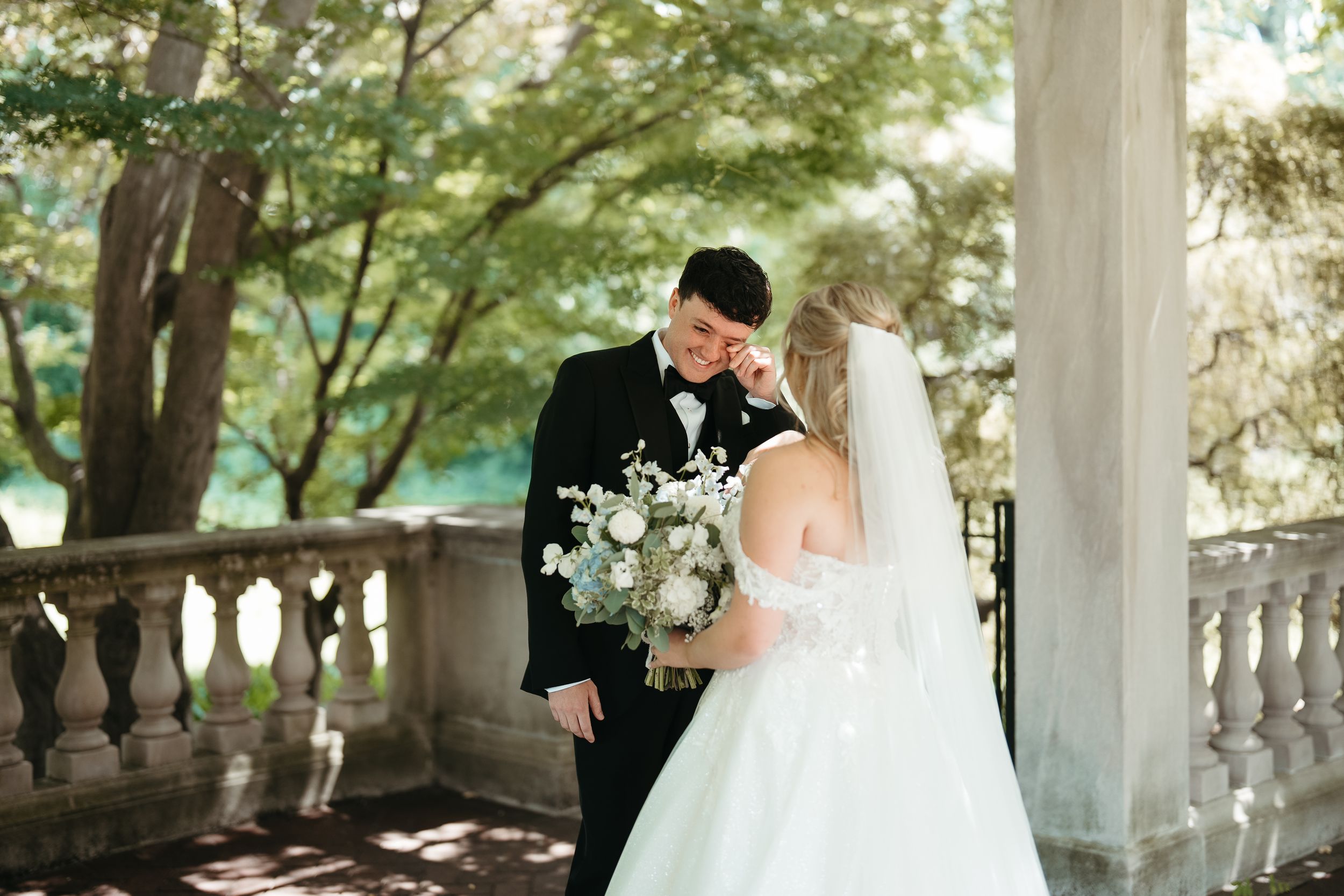 Bride and groom share a romantic moment on a stone balcony surrounded by lush green trees, bride holding white bouquet.