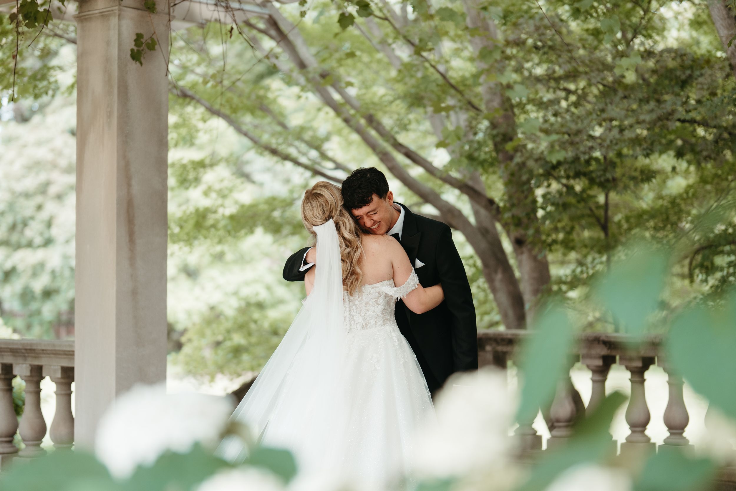 Bride and groom share a romantic embrace outdoors, surrounded by lush greenery and elegant white columns.