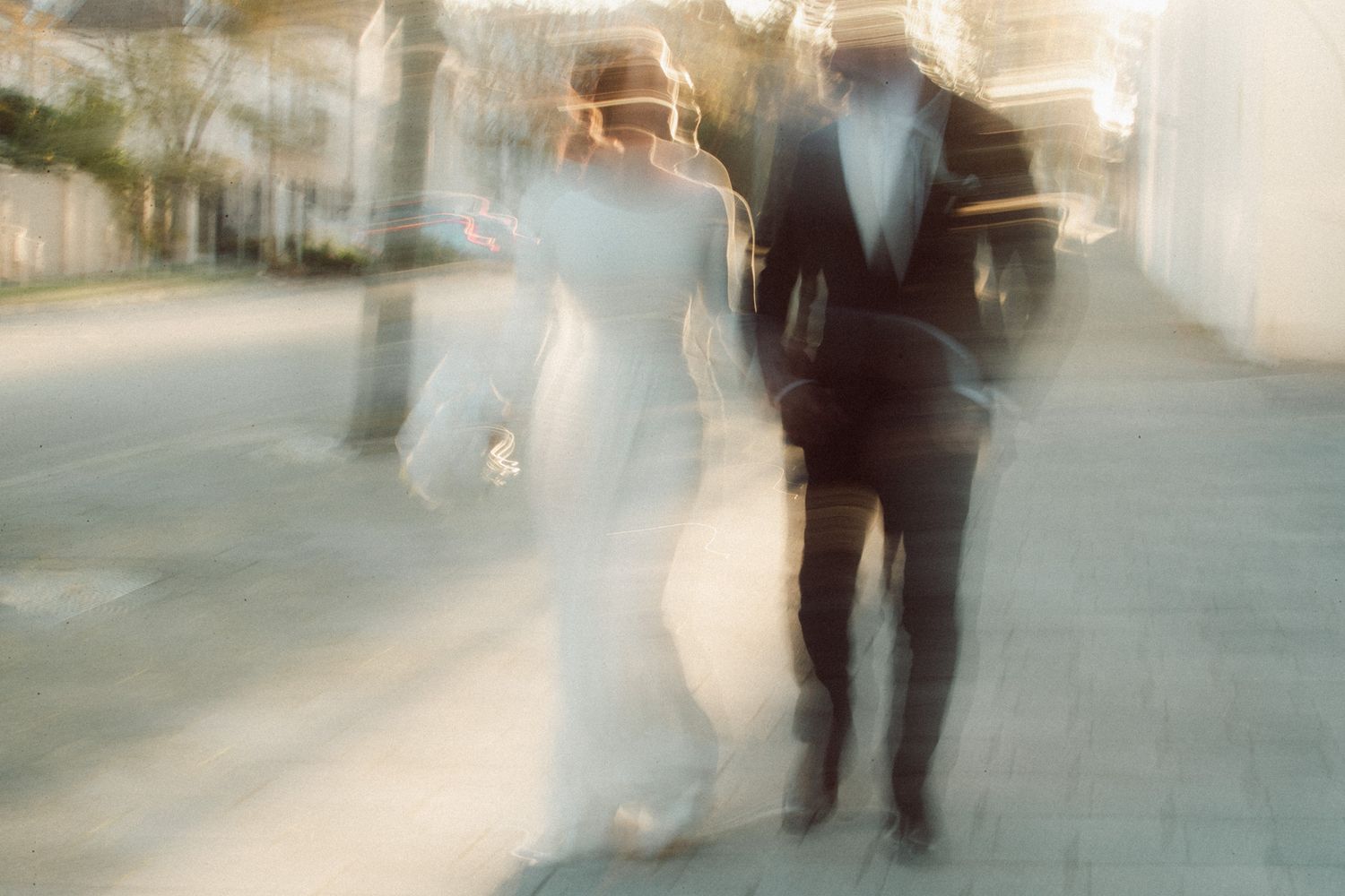 Blurry motion photo of bride in white gown and groom in black tuxedo walking together outdoors on sunny day.