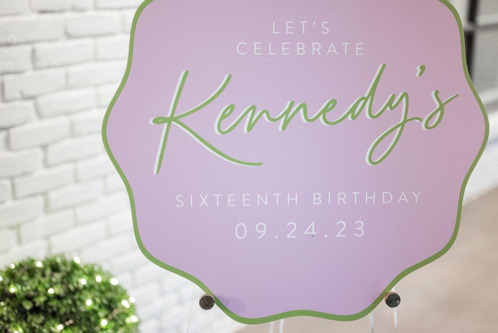 A lilac and mint green birthday sign reads 'Let's Celebrate Kennedy's Sixteenth Birthday' against a white brick wall.