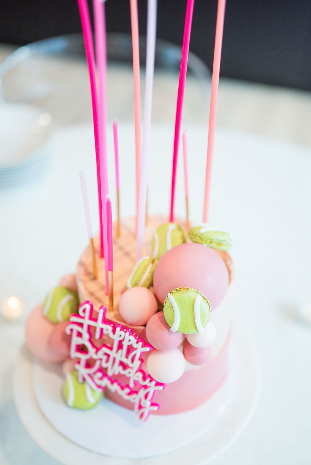 Pink birthday cake topped with tennis ball macarons and bright pink candles.