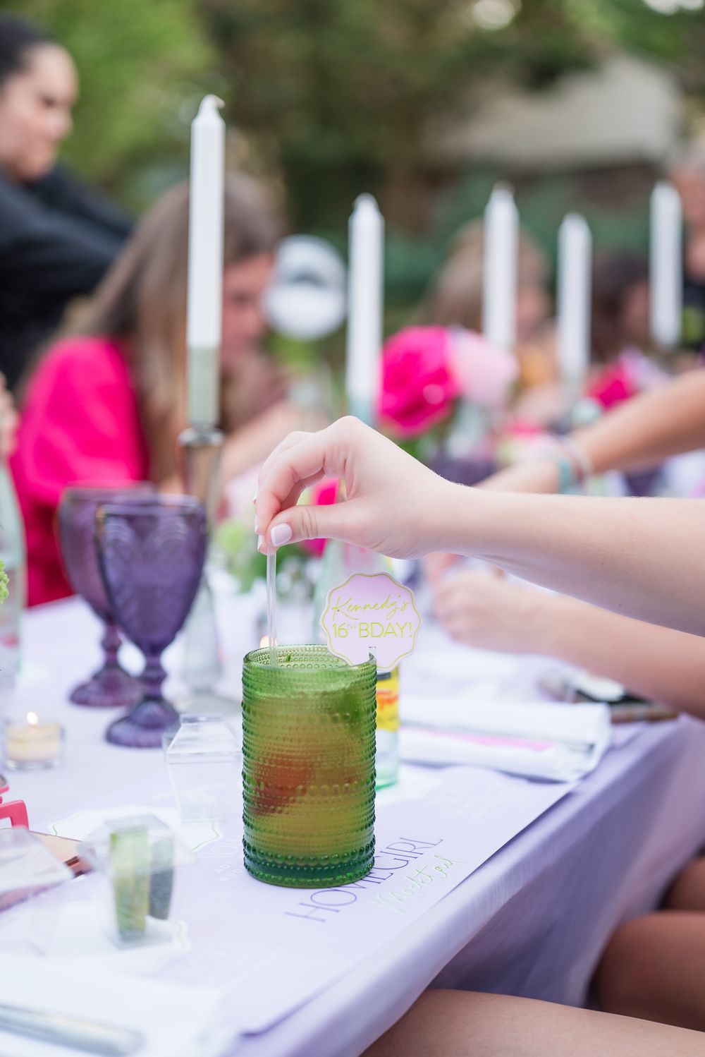A hand stirs a green drink in a glass at an elegant outdoor table setting with purple goblets and white candles.
