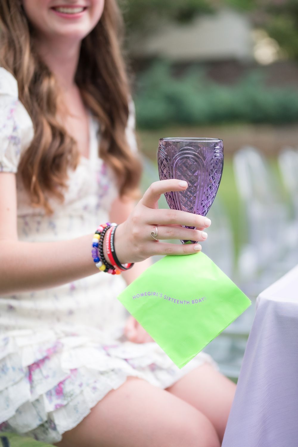 Person in white dress holding purple glass and green napkin at outdoor party.