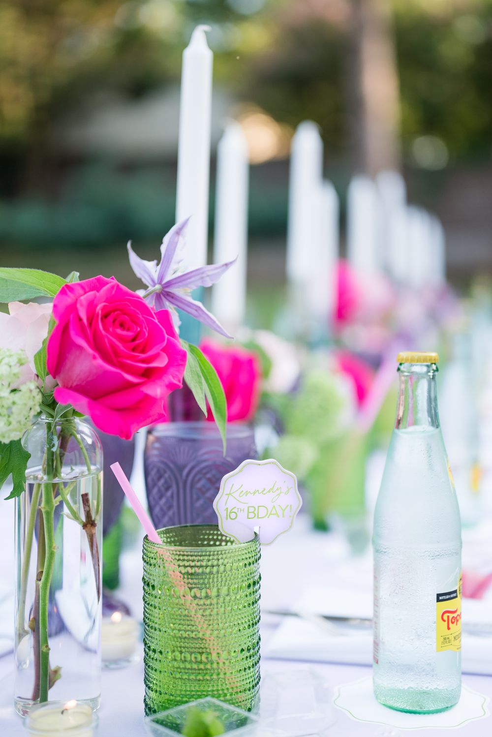 Table decoration with pink roses and purple glassware for outdoor celebration.