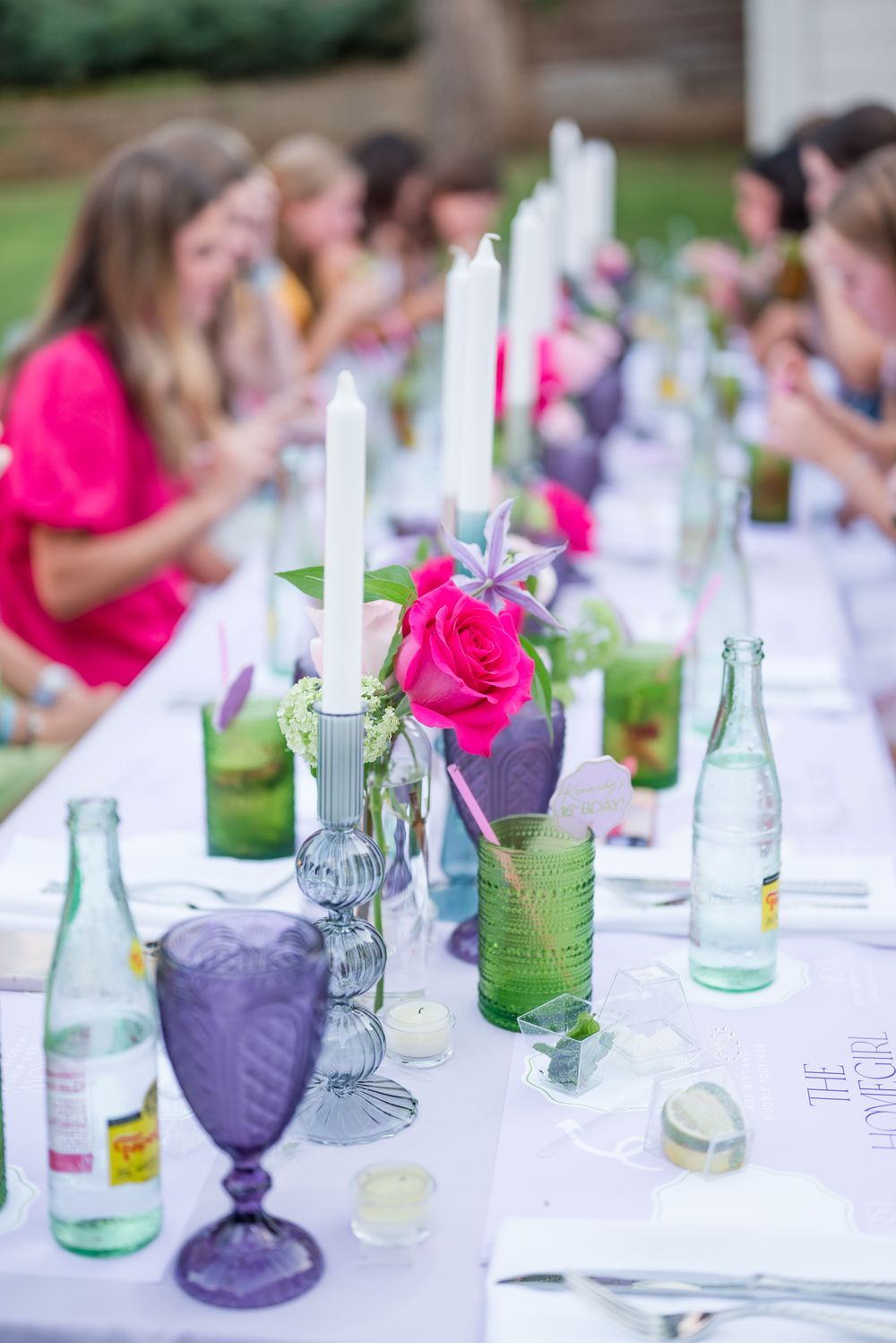 Elegant table setting with pink roses, purple glassware, white candles and green drinks at an outdoor party.