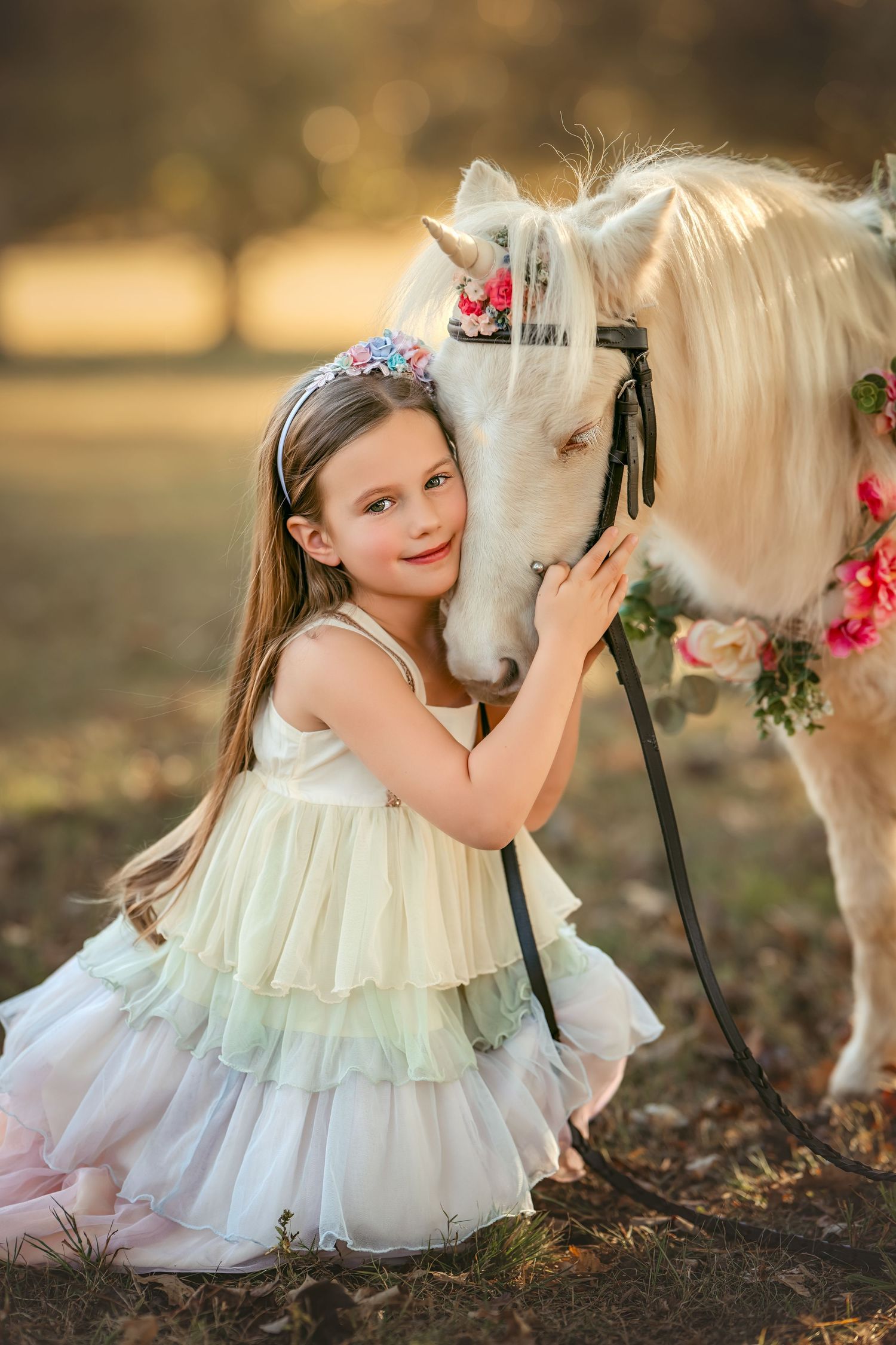 A young girl in a pastel rainbow dress embraces a white pony decorated with flowers in a sunlit field.