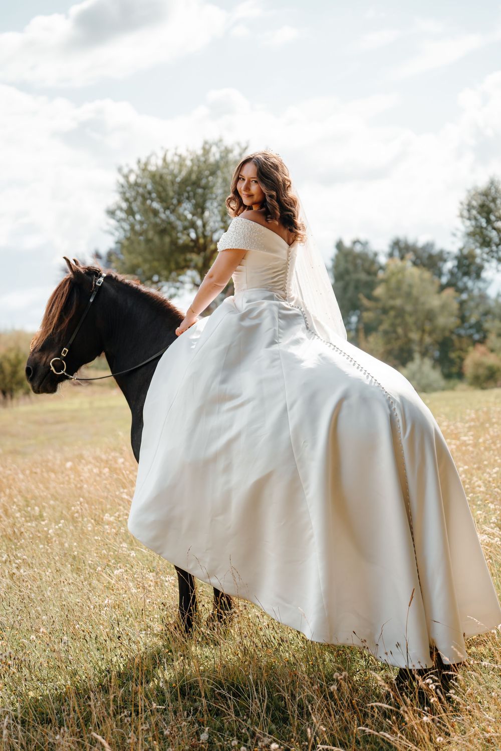 Bride in white gown and veil riding a dark horse in a sunny field with trees in the background.