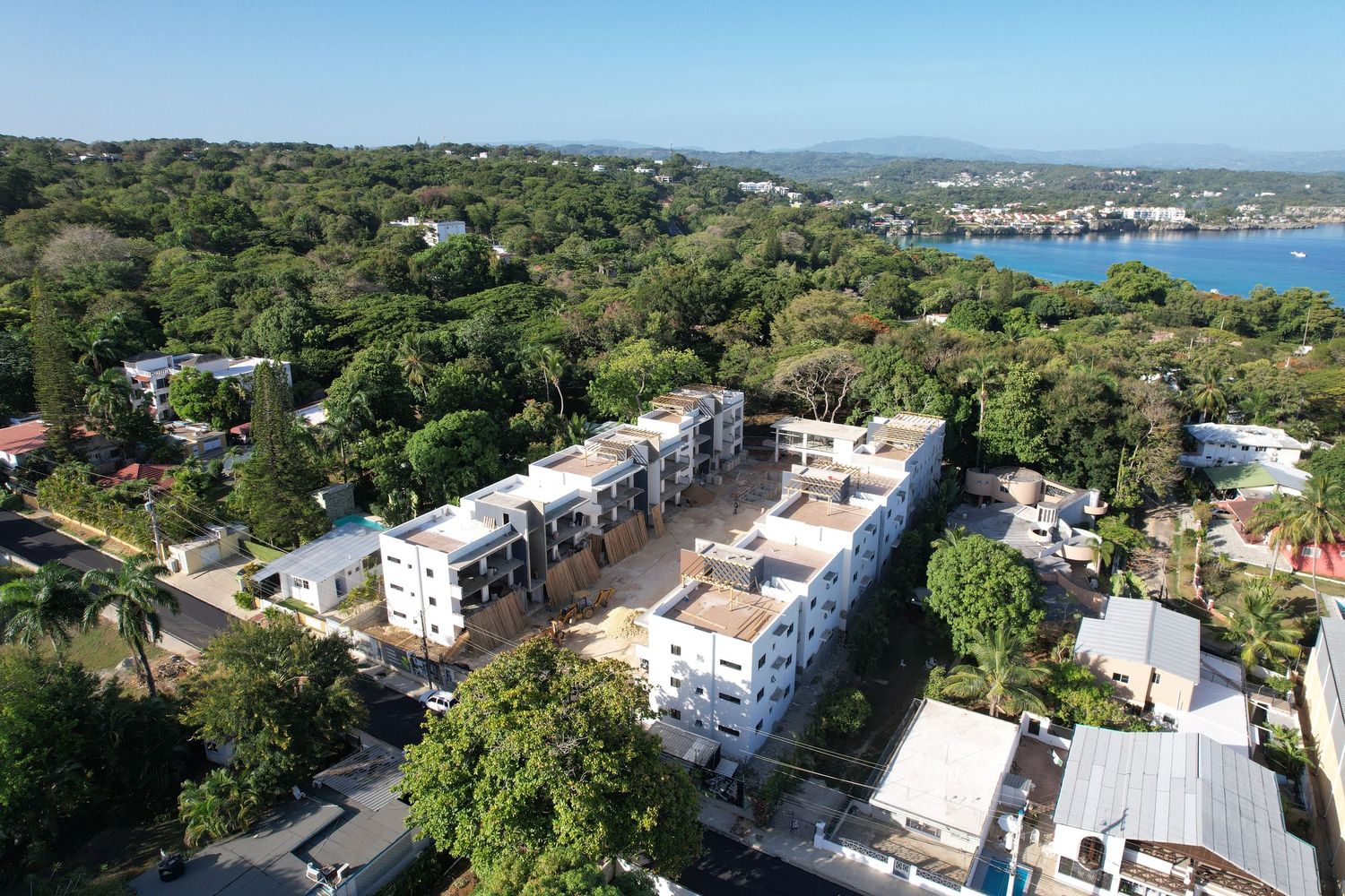 Aerial view of white multi-story residential complex surrounded by lush tropical trees near a coastal bay.