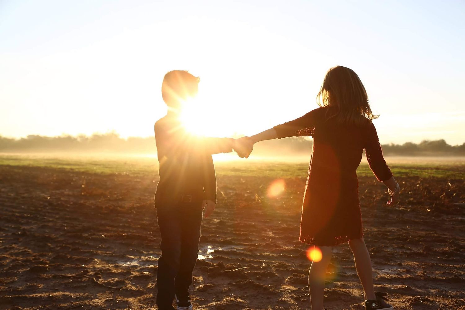 Two silhouetted children hold hands in a field at sunset, with bright golden sunlight and lens flare behind them.